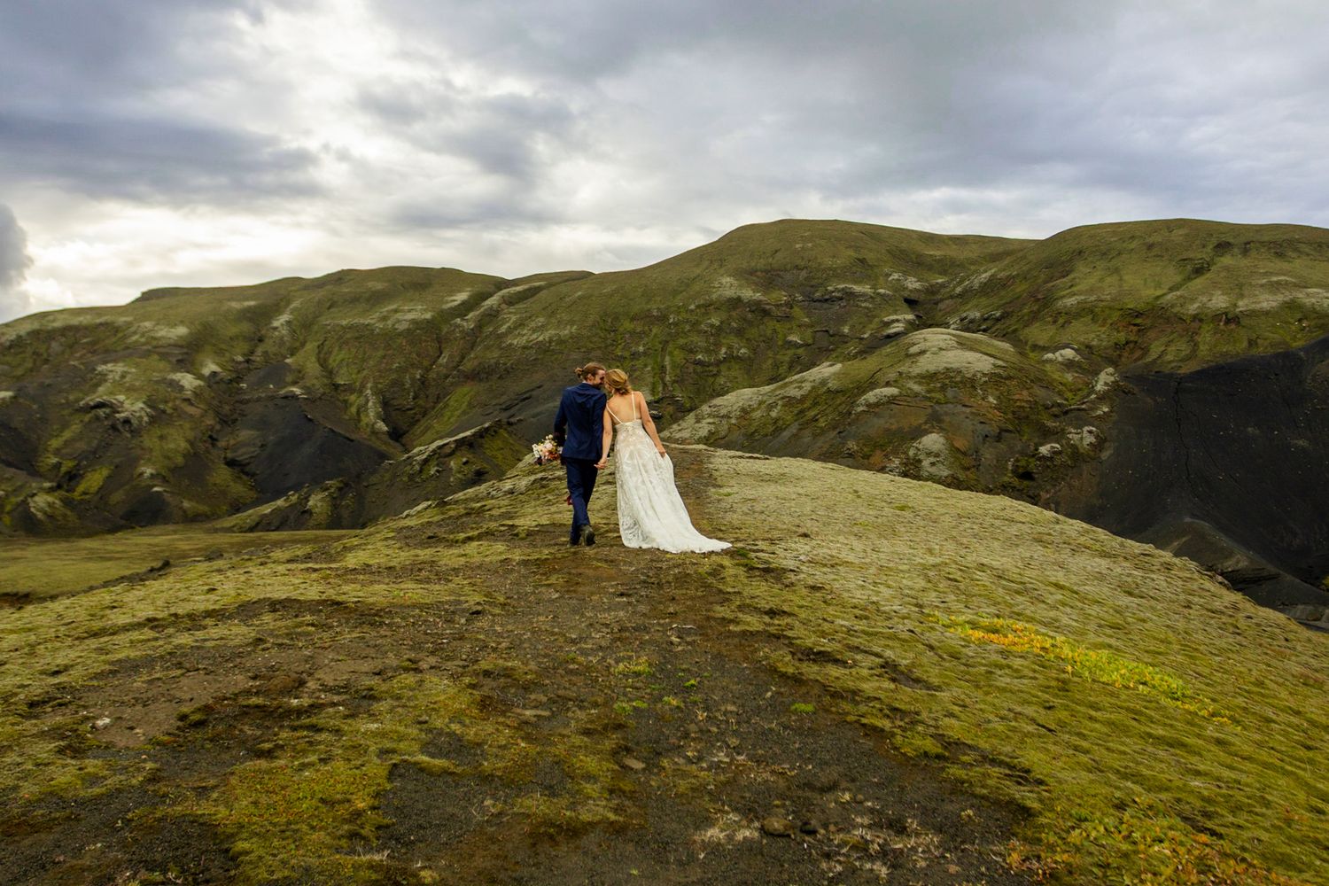 A couple stands atop a verdant mountain ridge under moody skies in Iceland.