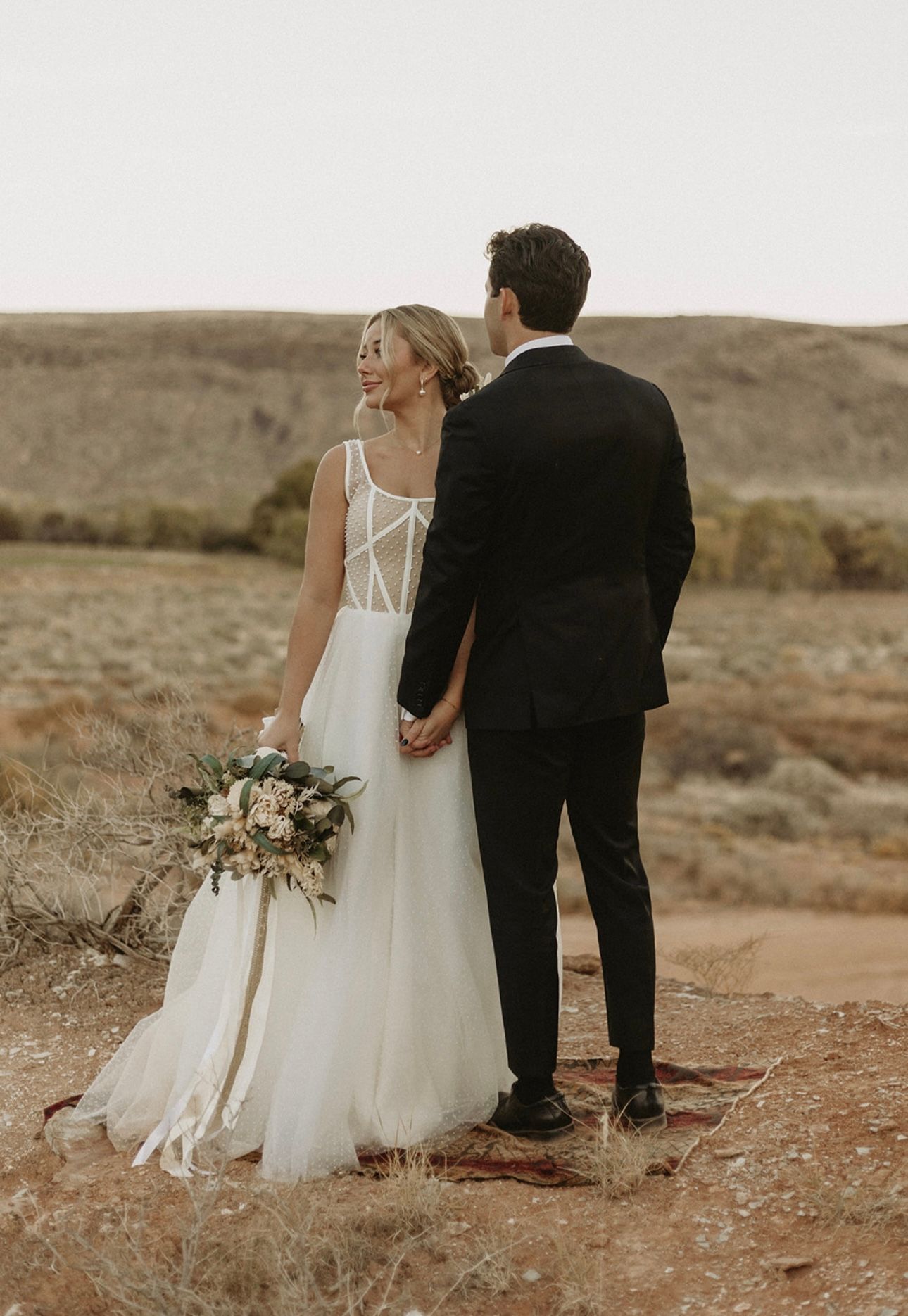 Couple in wedding attire holding hands while standing on desert terrain at sunset.