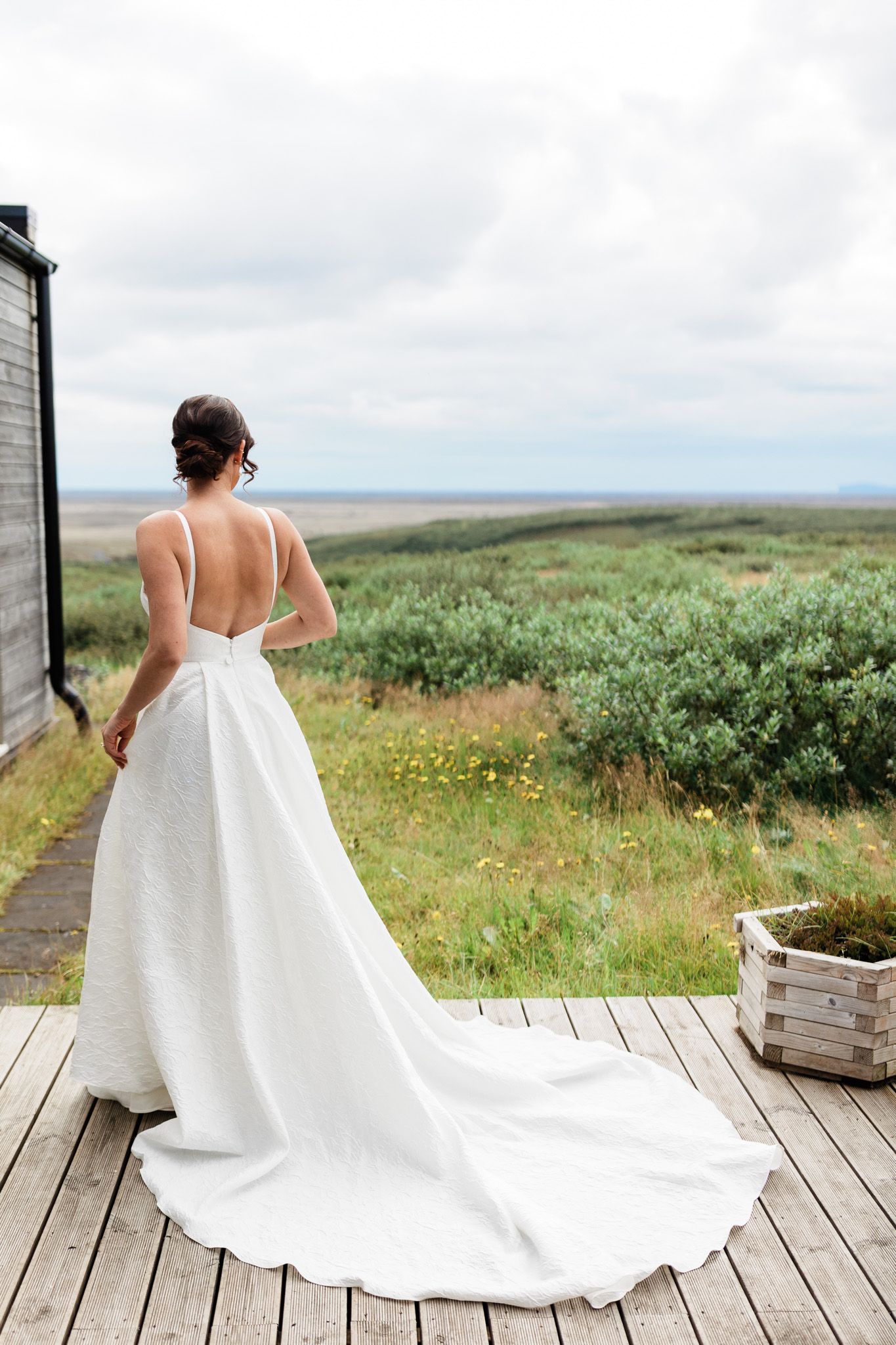 A bride in a flowing white wedding dress with a long train stands on a wooden deck overlooking coastal scenery.