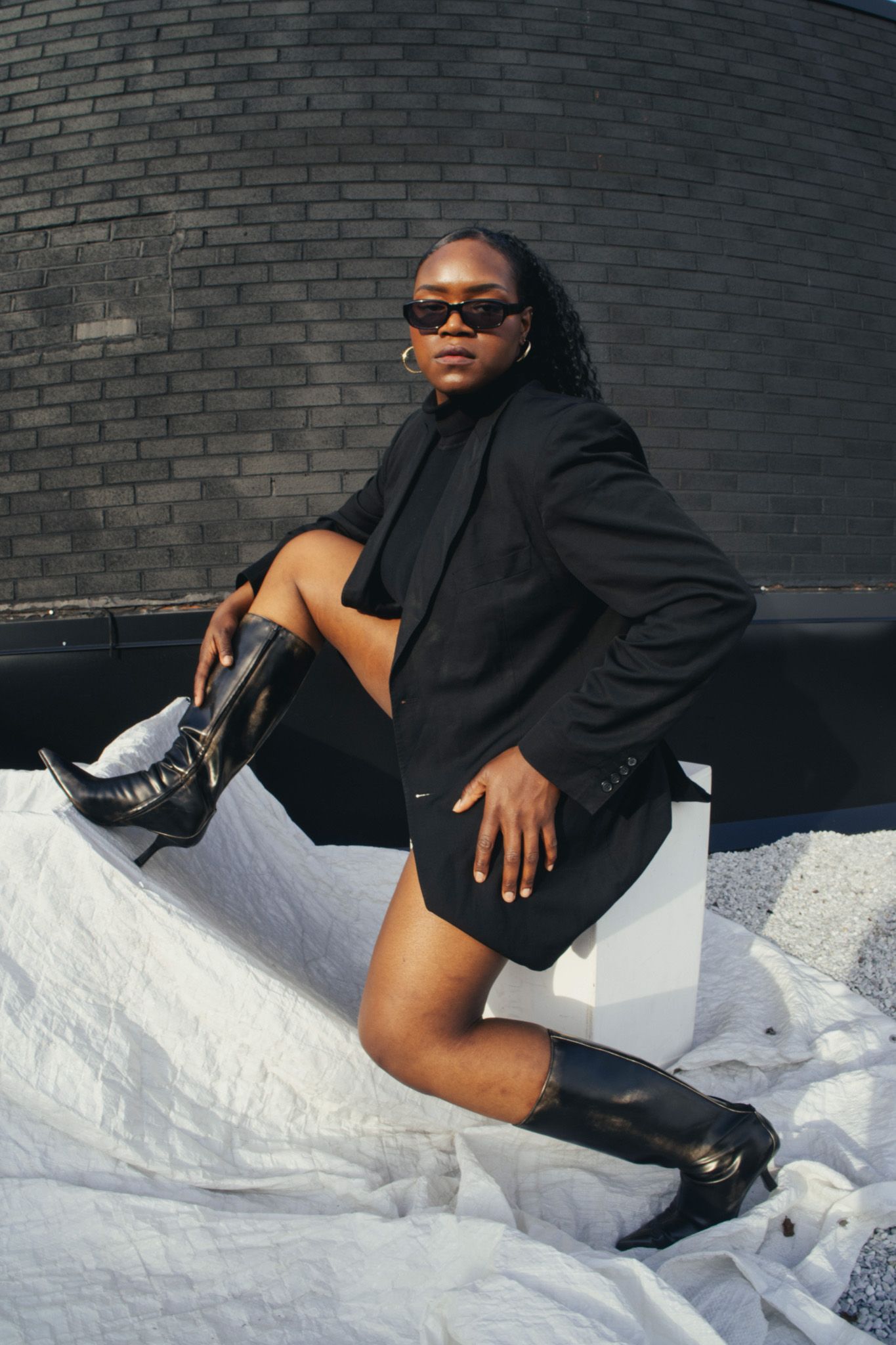 Fashion portrait of woman in black blazer and boots posing against brick wall.