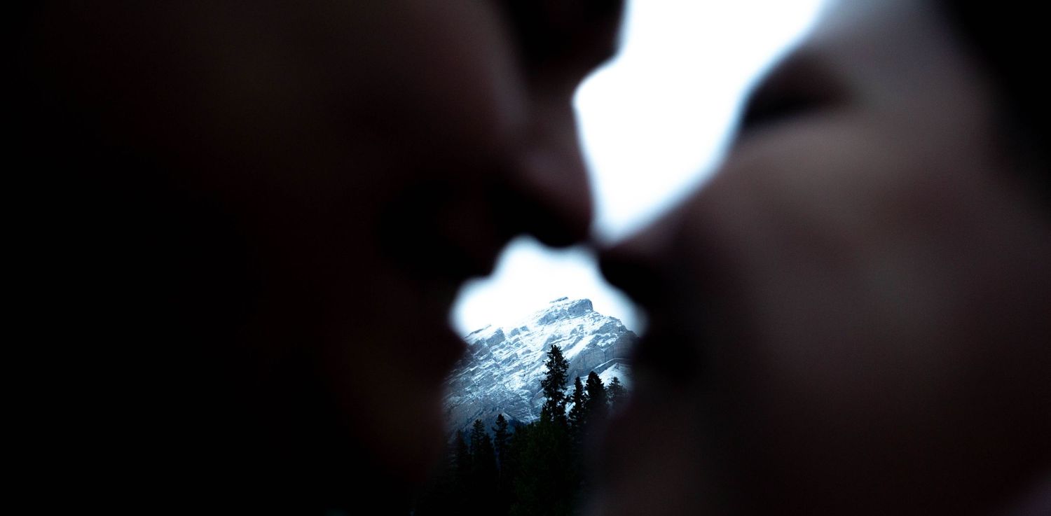 Silhouetted couple sharing an intimate moment with snowy mountain peaks visible between them.
