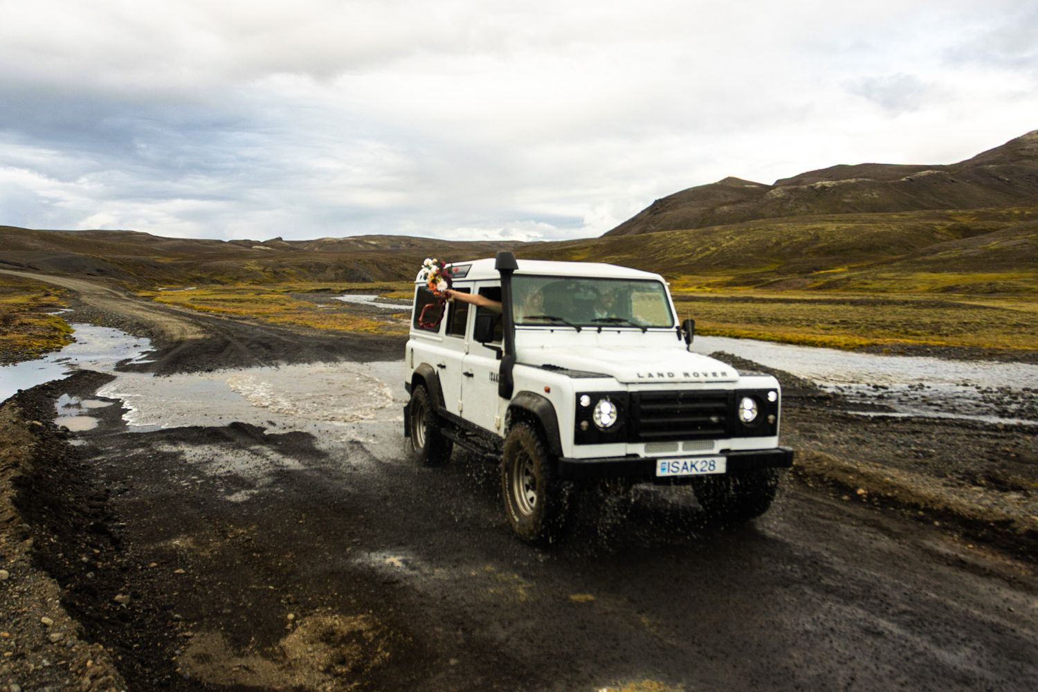 White Land Rover Defender driving through rugged mountain terrain in cloudy weather.