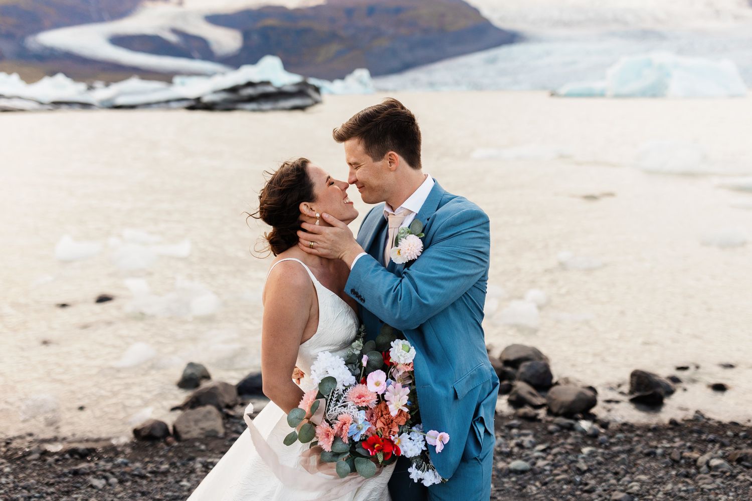 A wedding couple embraces on a rocky beach with glaciers and ice in the background in Iceland.