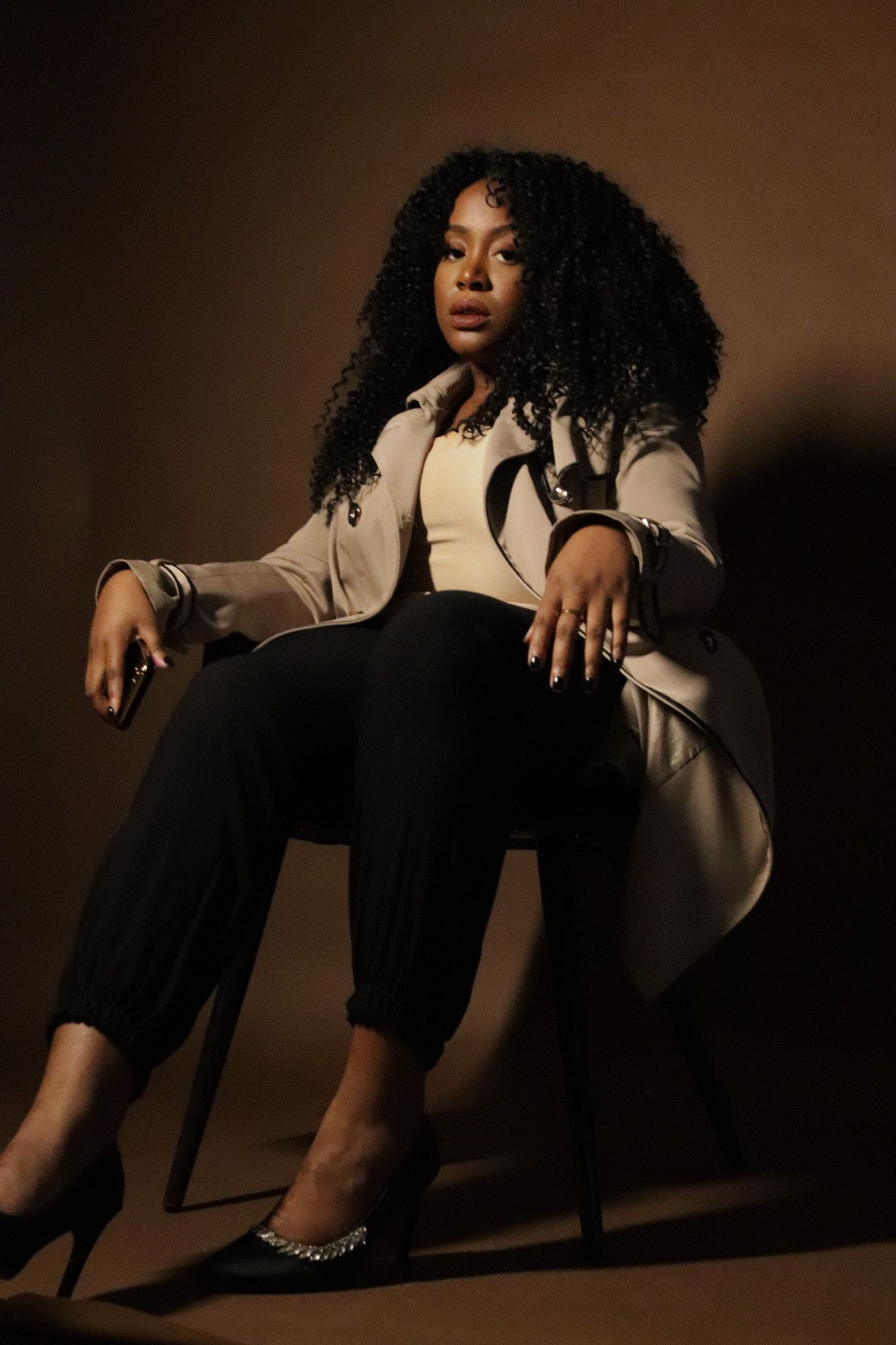 Professional portrait of person in beige blazer and black pants sitting on chair against brown background.