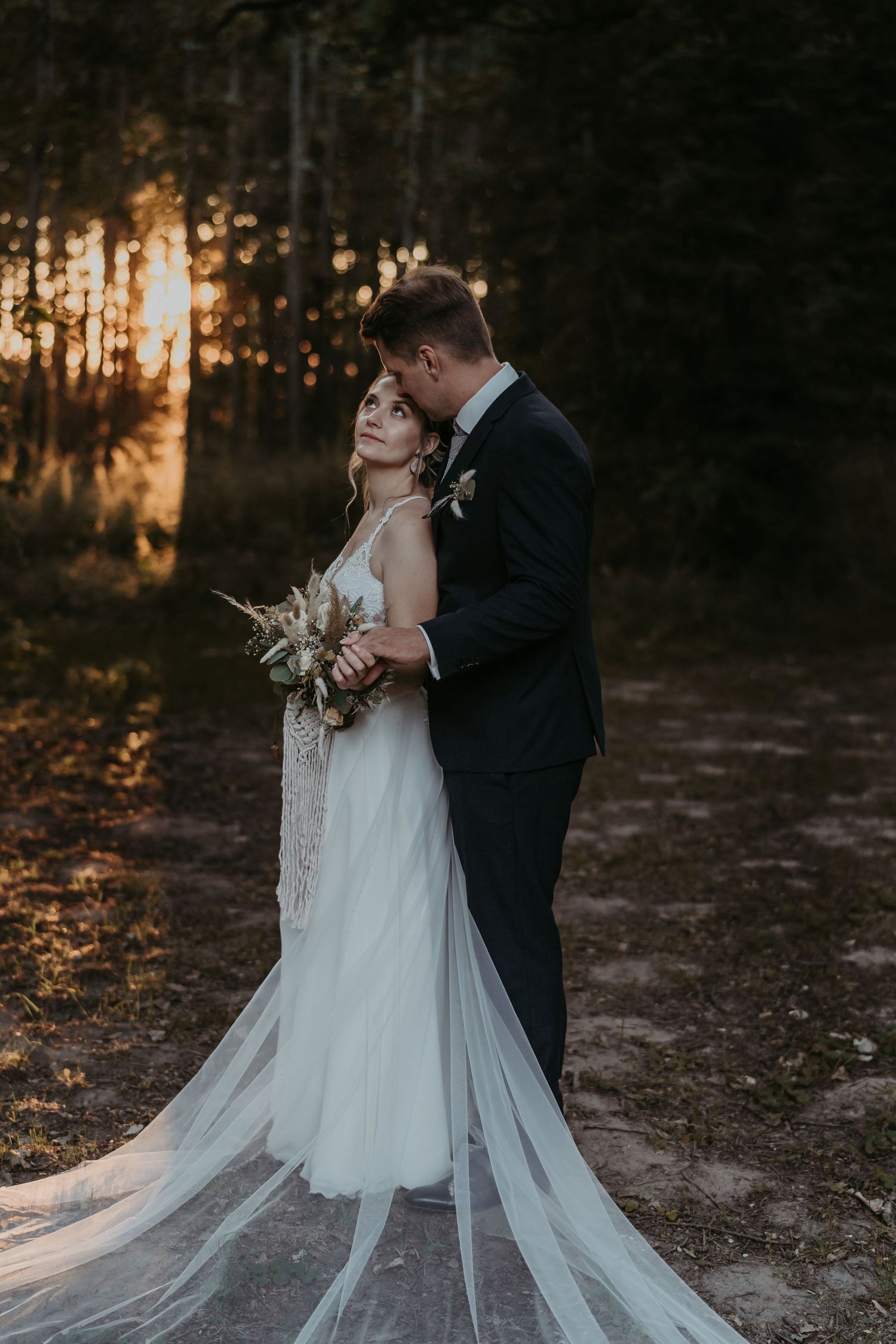 A couple embraces in a dreamy forest setting at sunset with a flowing white wedding dress and romantic lighting.