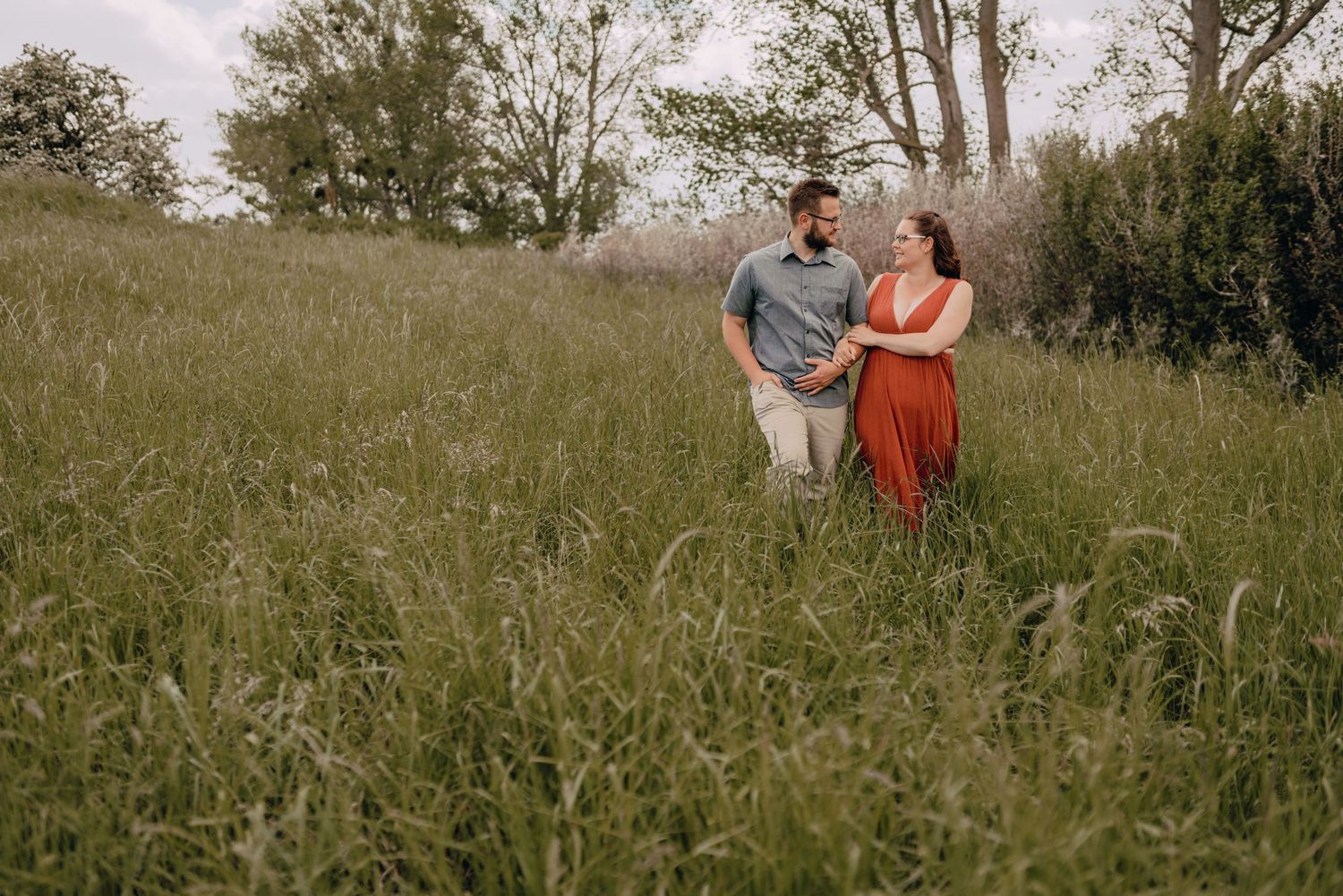 Couple stands together in pastoral field with trees in background during golden hour photoshoot.