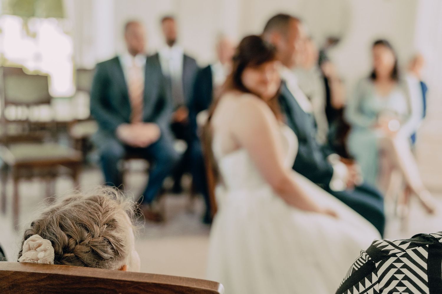 A young child observes a wedding ceremony from a wooden chair in the foreground.