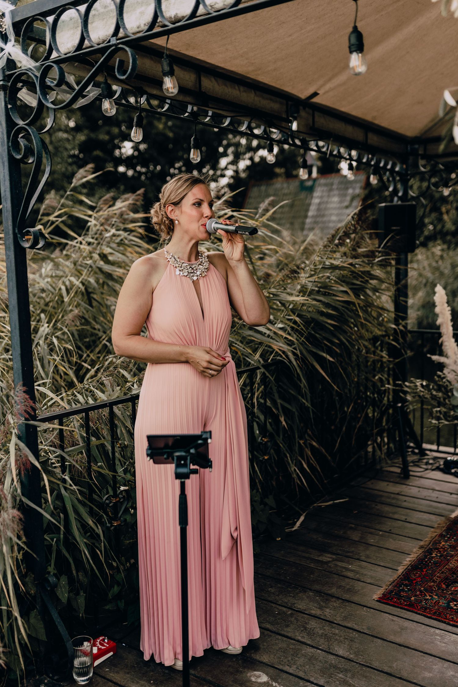 Elegant figure in flowing blush pink pleated gown stands on decorated patio with string lights and ornamental grasses.