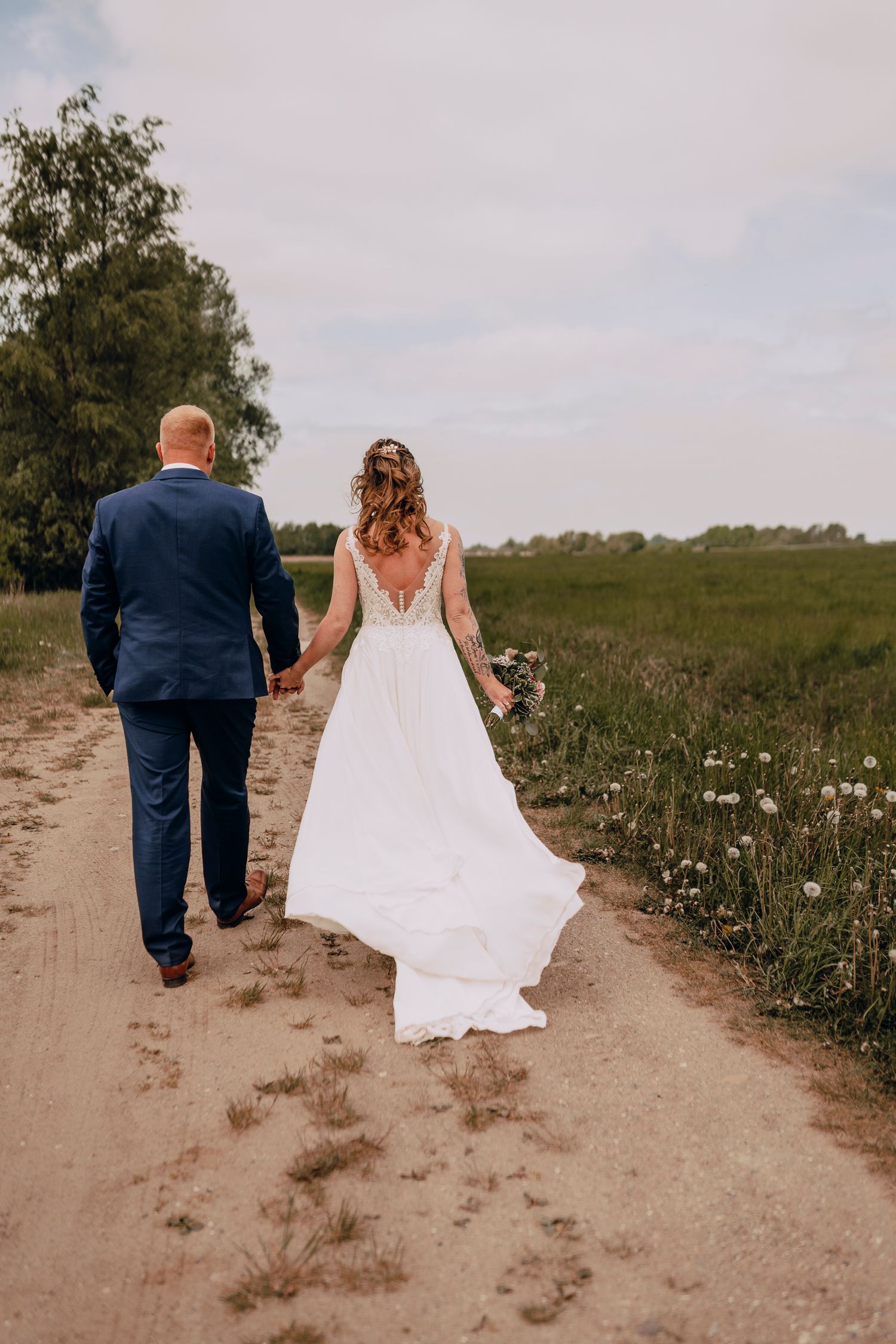 Newlyweds walk along a rustic dirt path with wildflowers and greenery in the background.