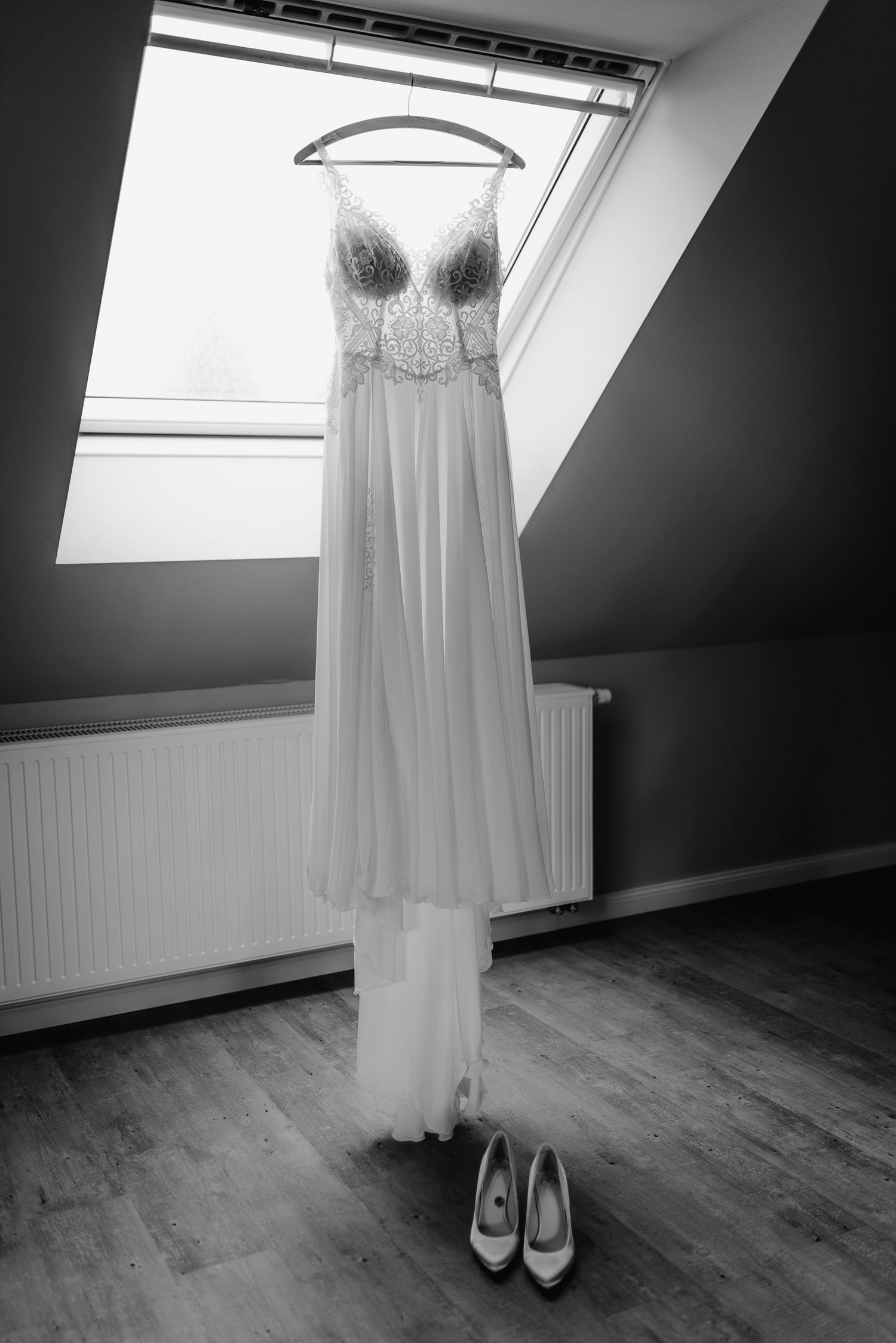 A delicate white bridal dress and silver shoes displayed in a minimalist attic room with sloped ceiling and wooden floors.