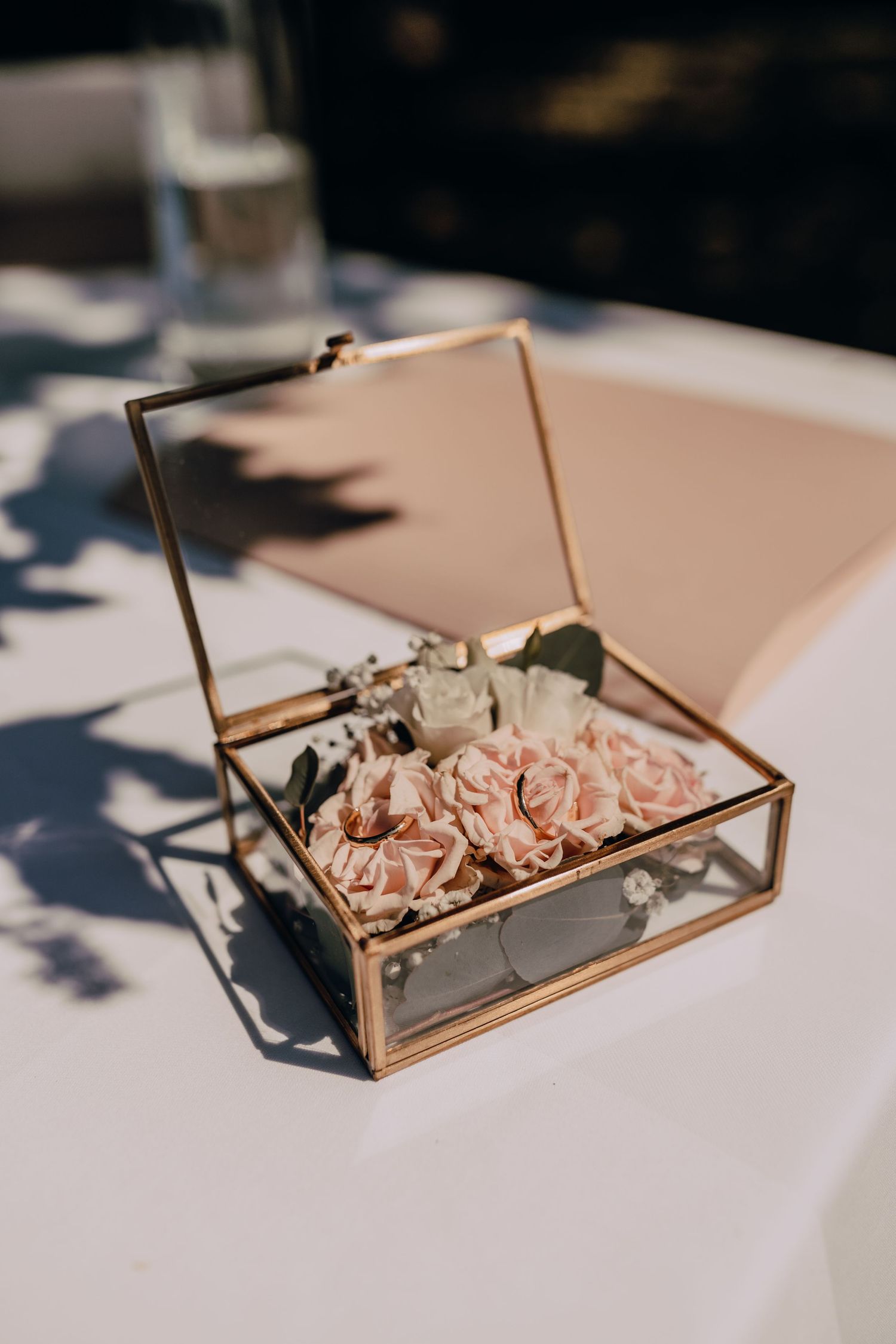 Glass and brass jewelry box filled with delicate pink roses sitting on a white surface in natural light.