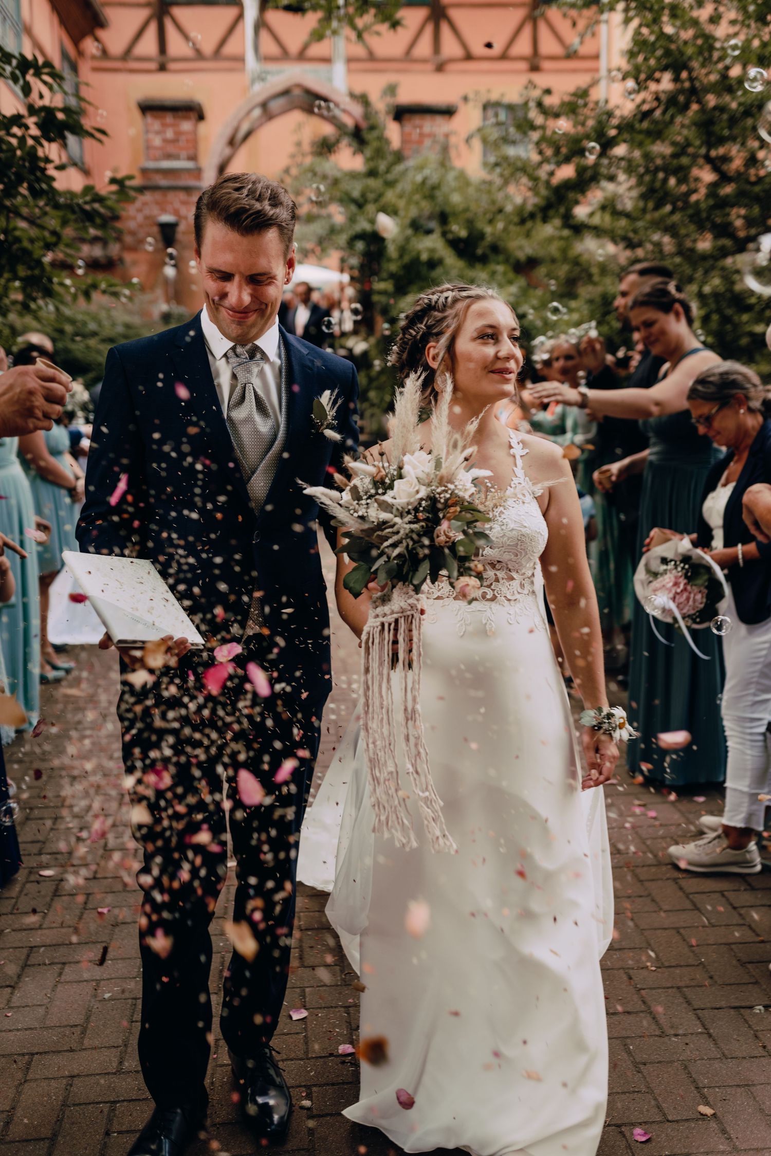 A wedding couple celebrates with guests throwing confetti outside a venue with coral-colored walls and brick pavers.