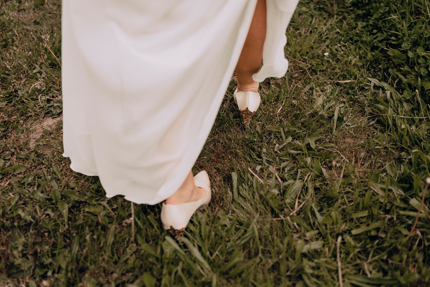 A person wearing a white wedding dress stands in grass wearing elegant cream-colored heels.