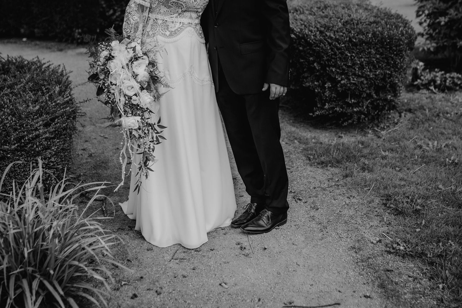 A black and white photograph shows the lower portion of a wedding couple on a garden path with ornamental grasses nearby.
