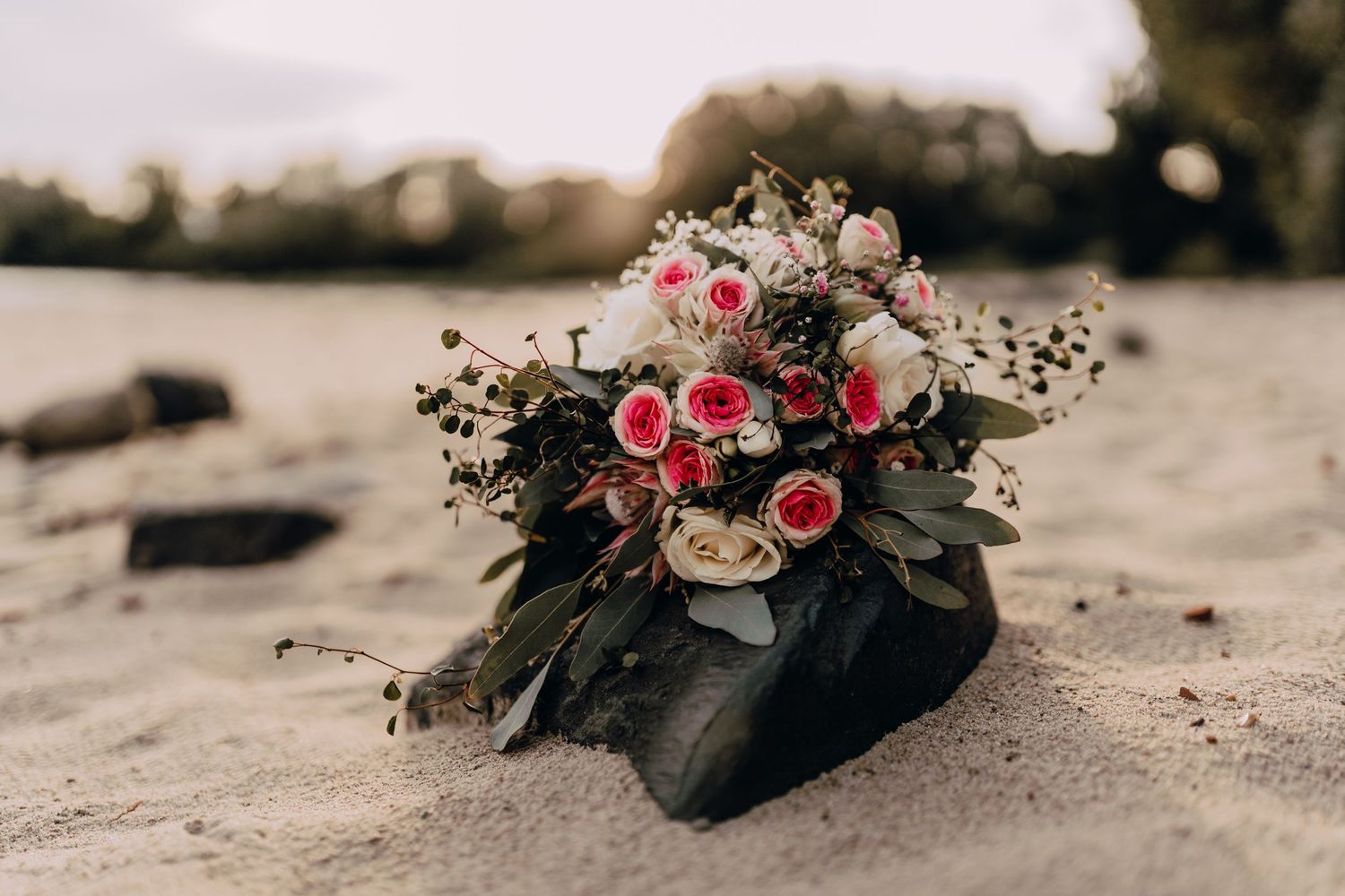 Romantic floral bouquet with pink roses and dark foliage lies on a sandy beach in vintage tones.