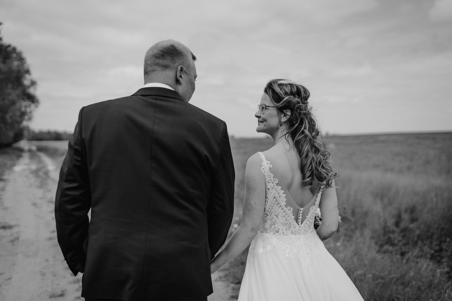 A black and white photo shows two people walking together in an open field during a wedding photo session.