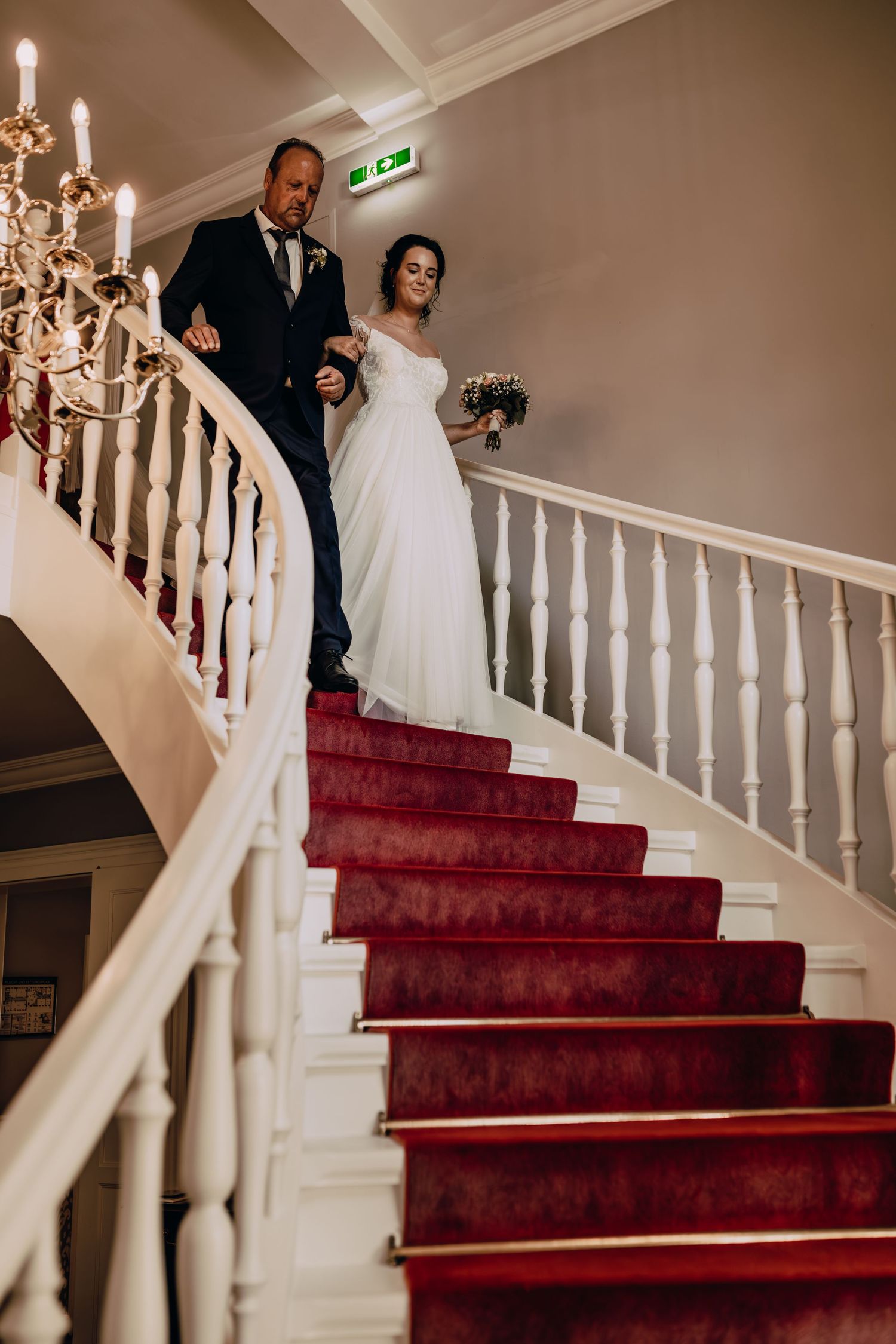 A formal couple descending an elegant curved staircase with a red carpet and white railings.