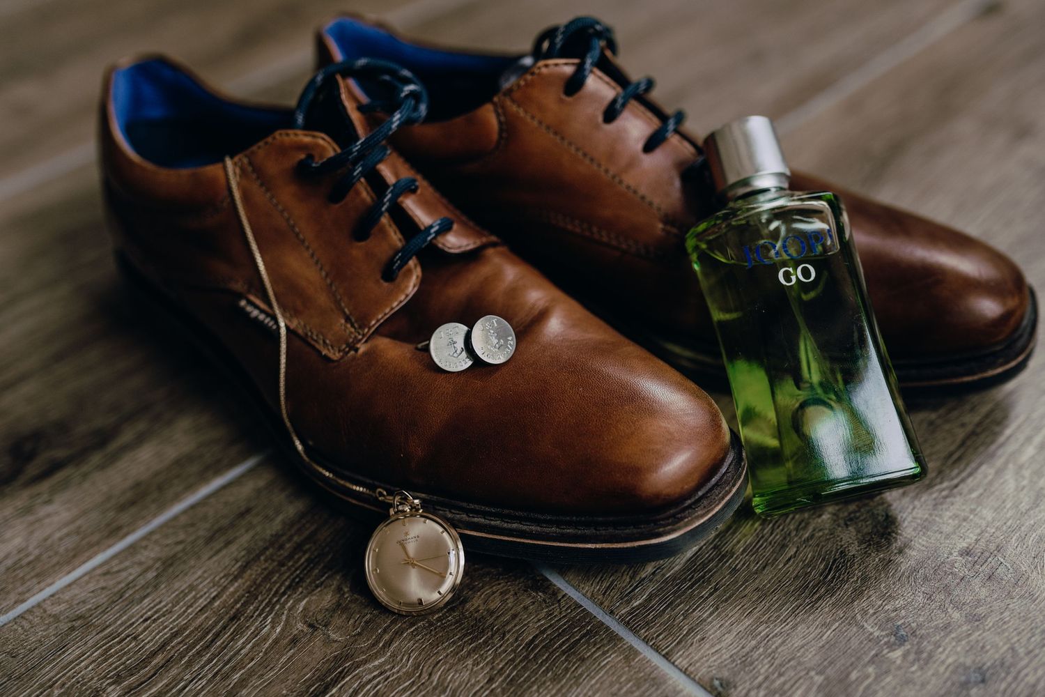 Brown leather dress shoes arranged with perfume bottles and greenery on wooden surface for wedding detail photography.