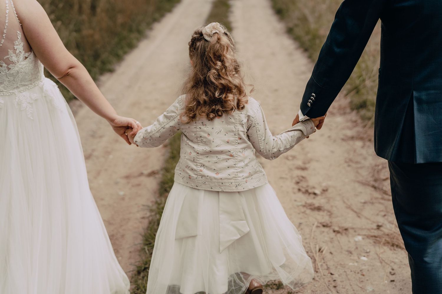 A small child in a white dress holds hands with bride and groom while walking down a country path.
