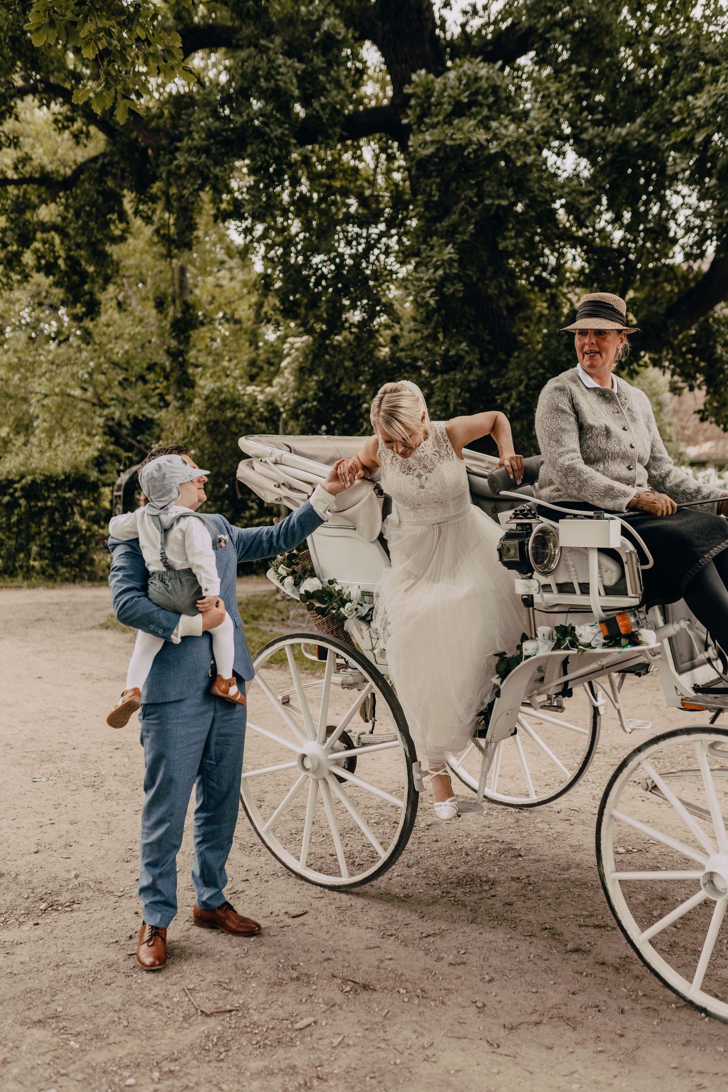 A wedding party gathers around an elegant white carriage on a tree-lined path during a rustic outdoor celebration.