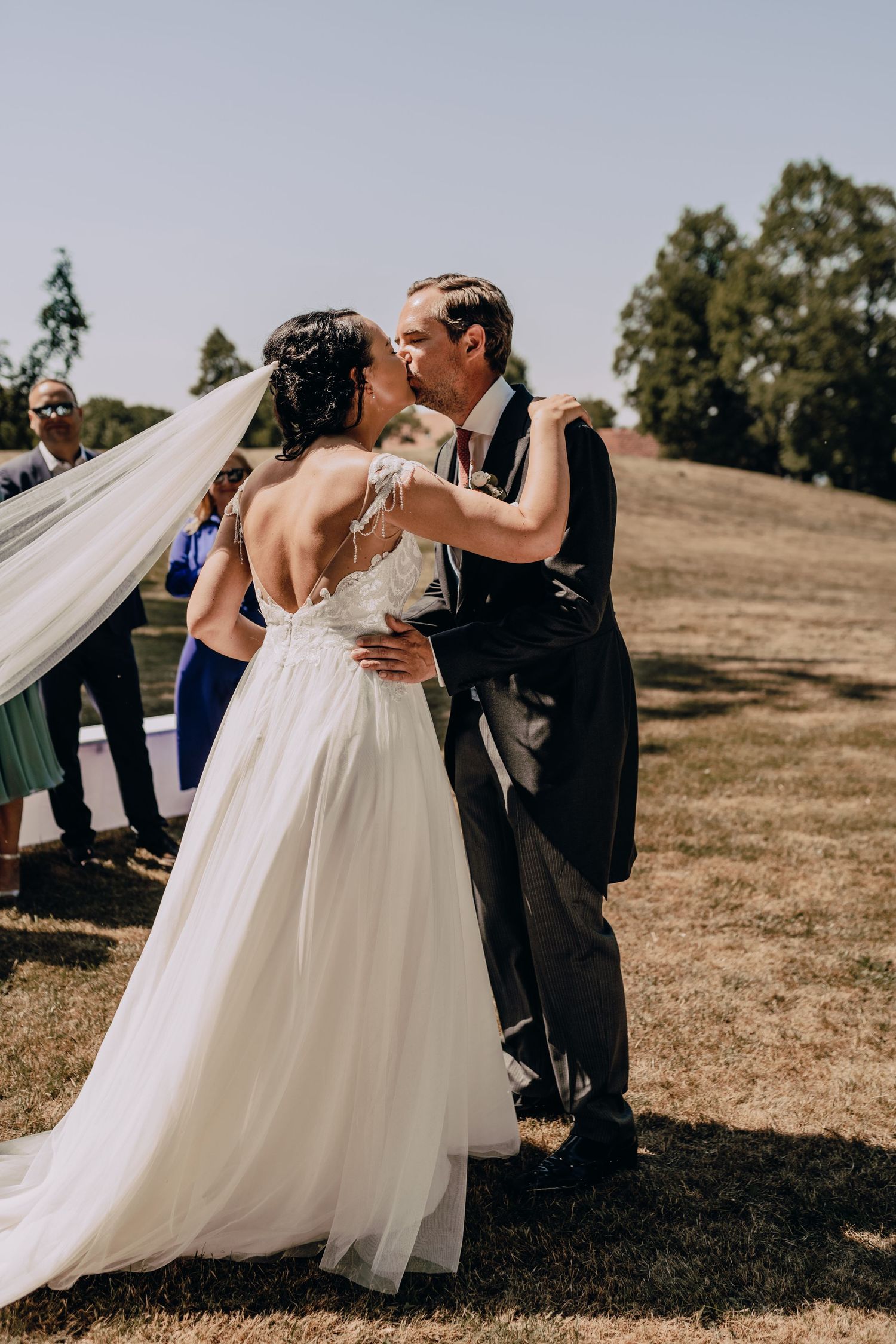 A bride and groom share their first wedding dance outdoors on a sunny day as her white veil flows in the breeze.
