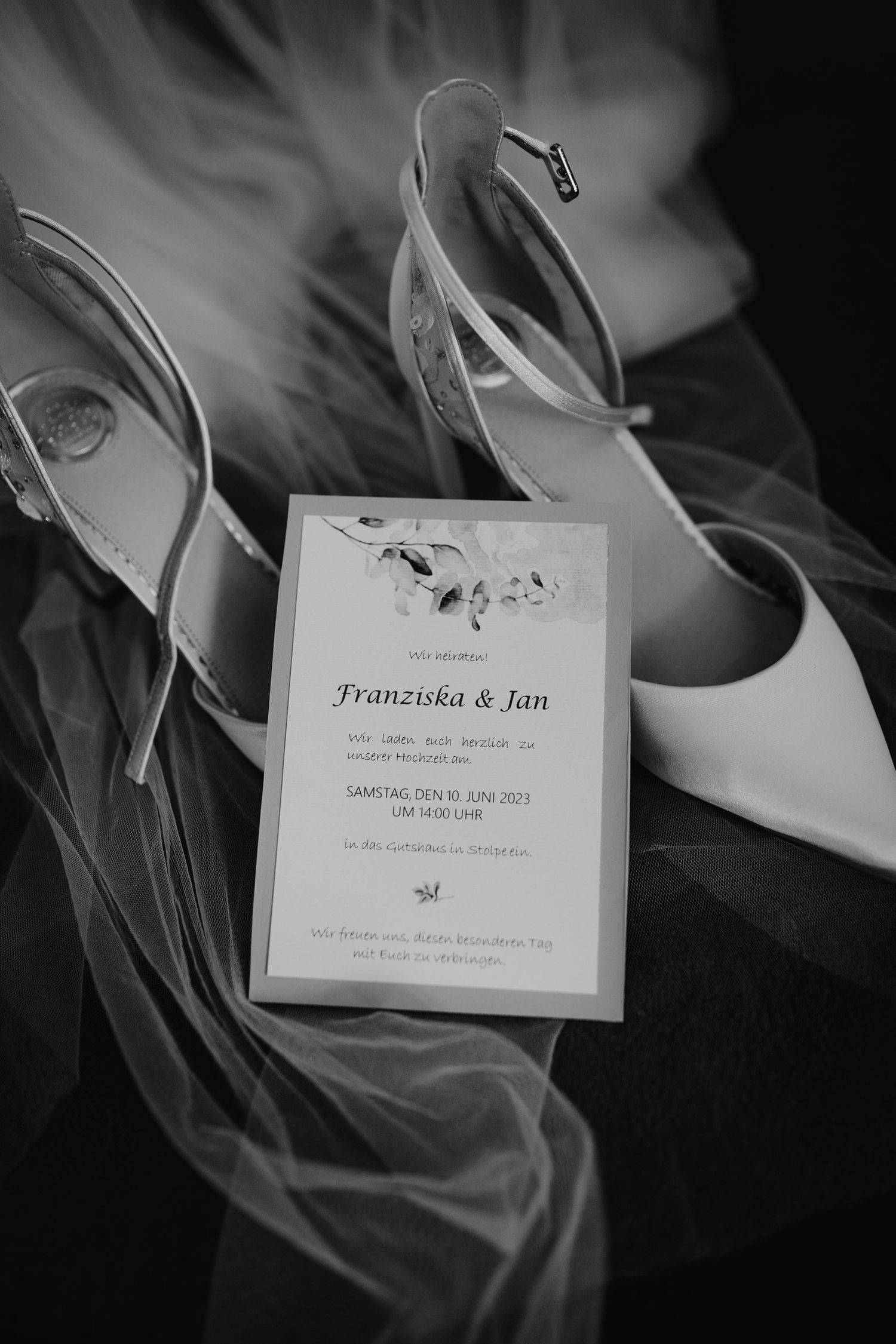 Wedding invitation resting on elegant white bridal shoes with a delicate veil backdrop in black and white.