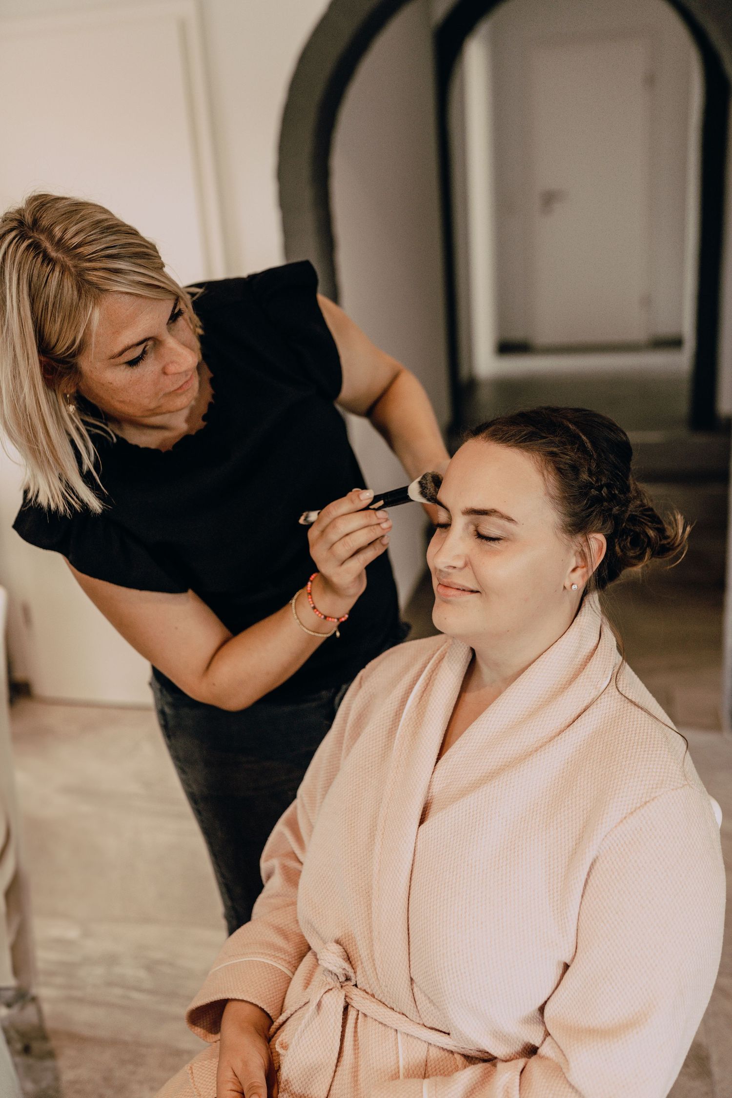 Makeup artist working on client wearing a blush pink robe during a beauty preparation session.