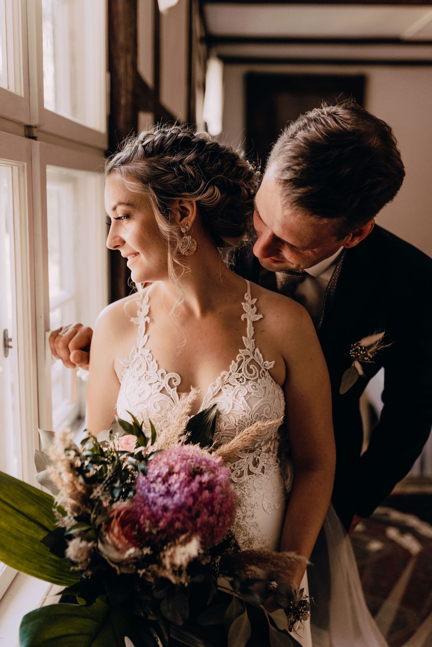 Intimate wedding moment by window with lace dress and colorful bouquet featuring pink and purple flowers.