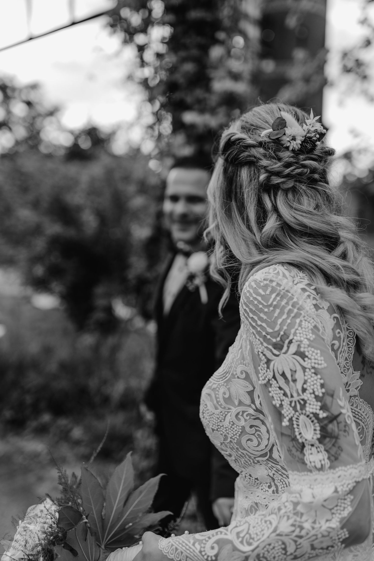 Black and white wedding photo showing bride in detailed lace dress with braided hair adorned with flowers.