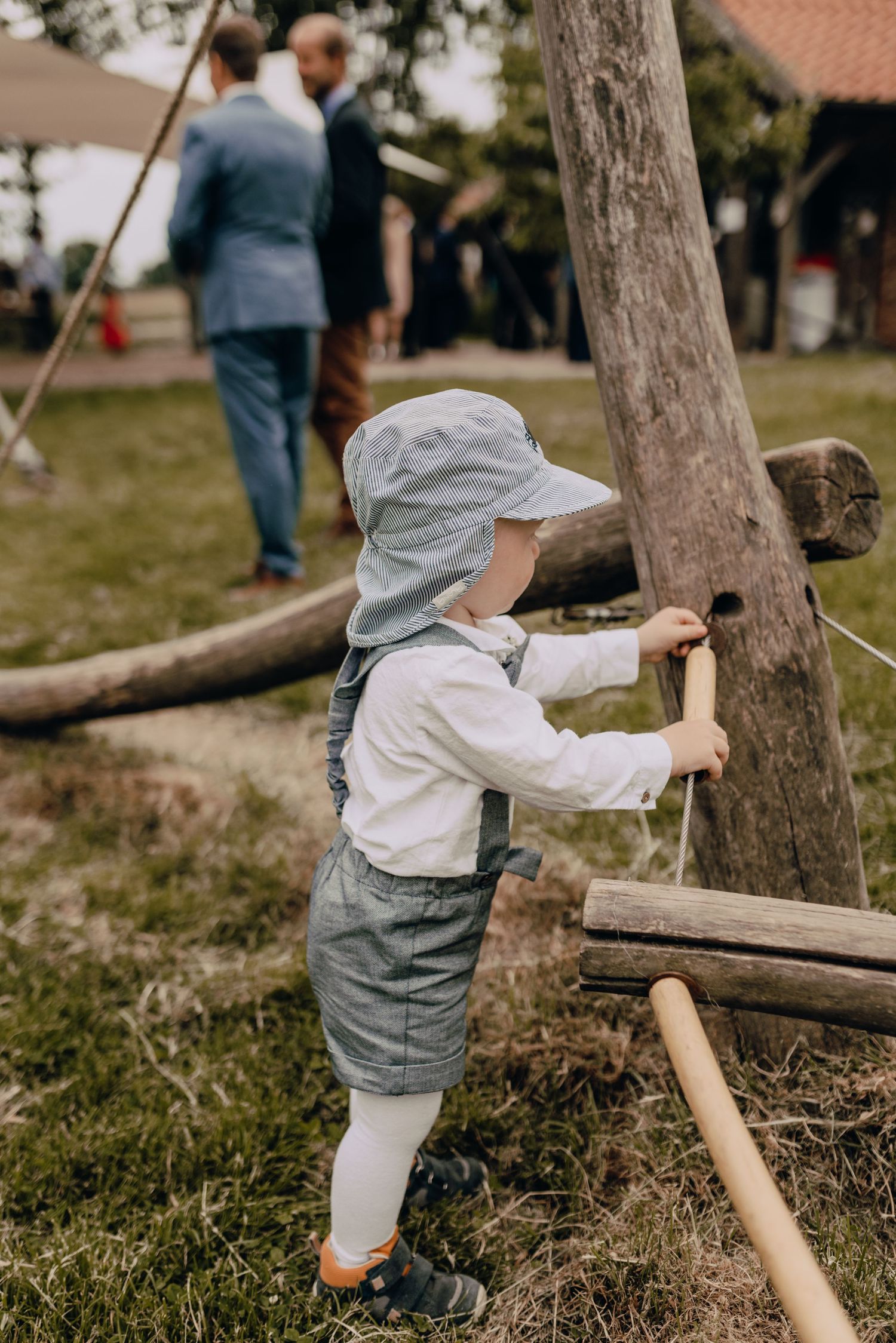 Small child in overalls explores outdoor natural play area using wooden sticks and poles near trees.