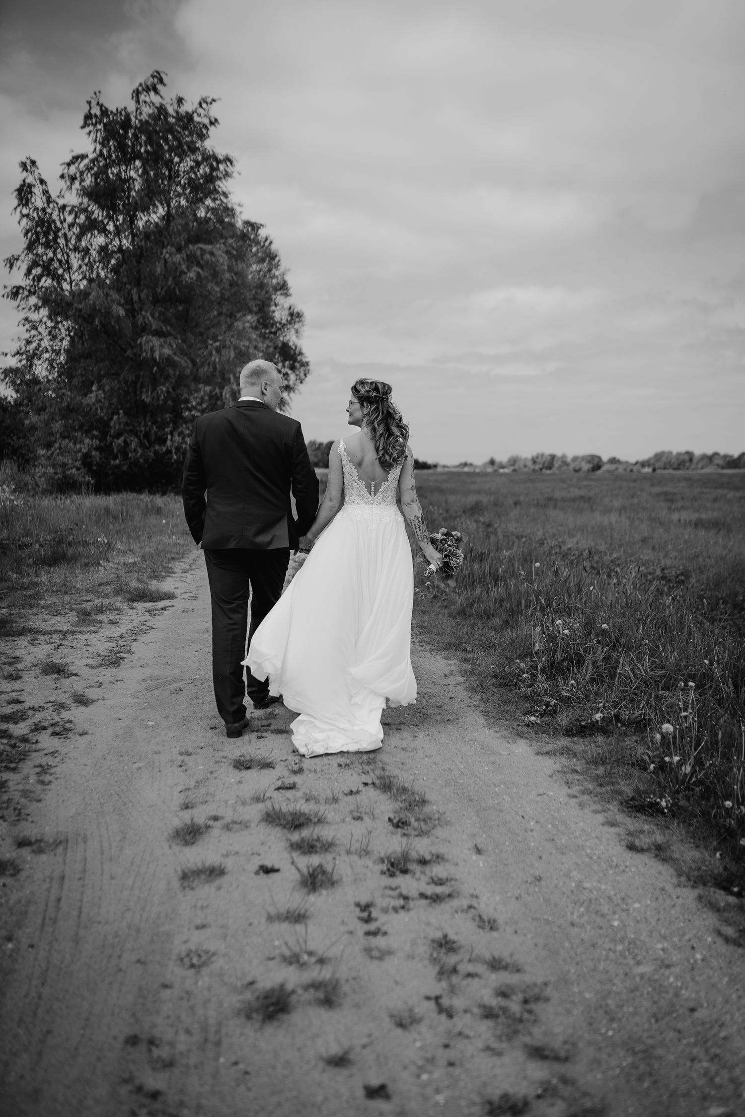 A newlywed couple walk hand in hand down a dirt path through an open field in a black and white photograph.