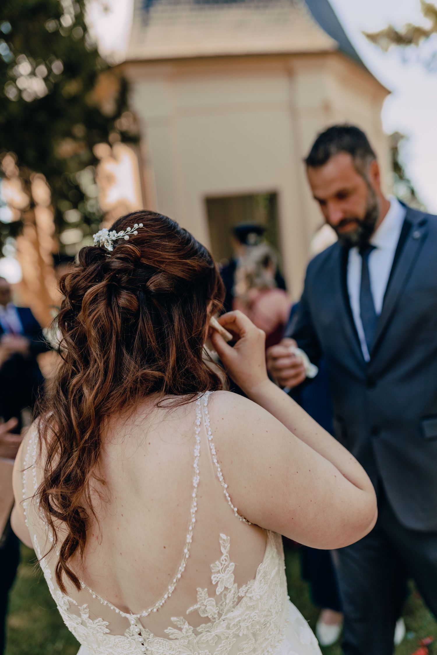 A bride in a lace wedding dress with open back stands with someone wearing a navy suit during an outdoor ceremony.