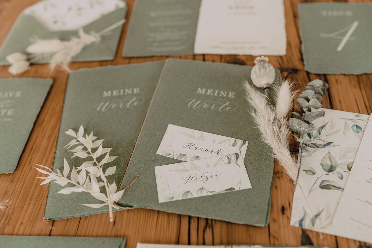 Sage green wedding stationery with botanical accents and calligraphy laid out on a wooden surface.