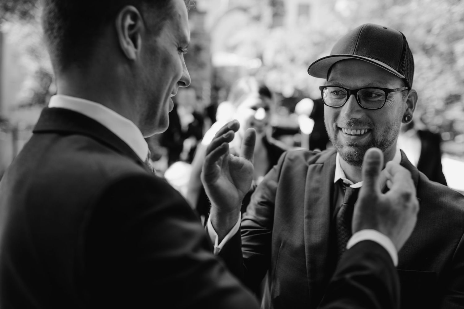 A black and white candid photo shows two people in conversation, with one wearing a suit and another in a baseball cap.