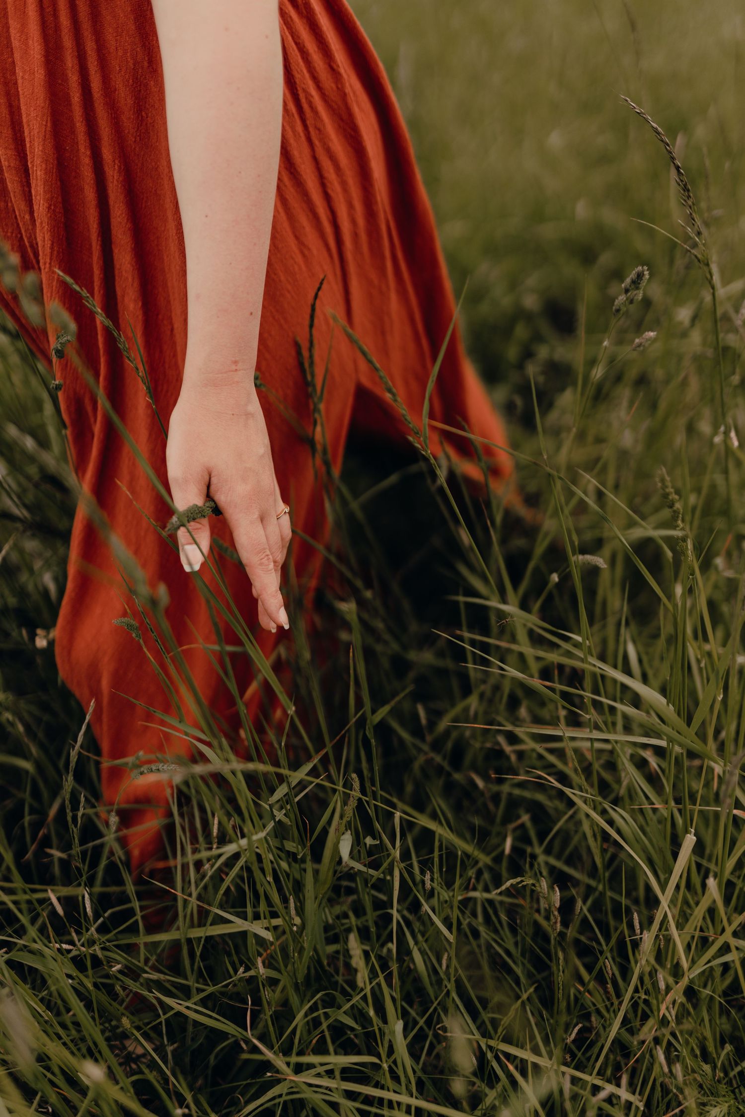 A hand wearing rings gracefully brushes against tall grass while wearing a flowing rust-colored dress in nature.