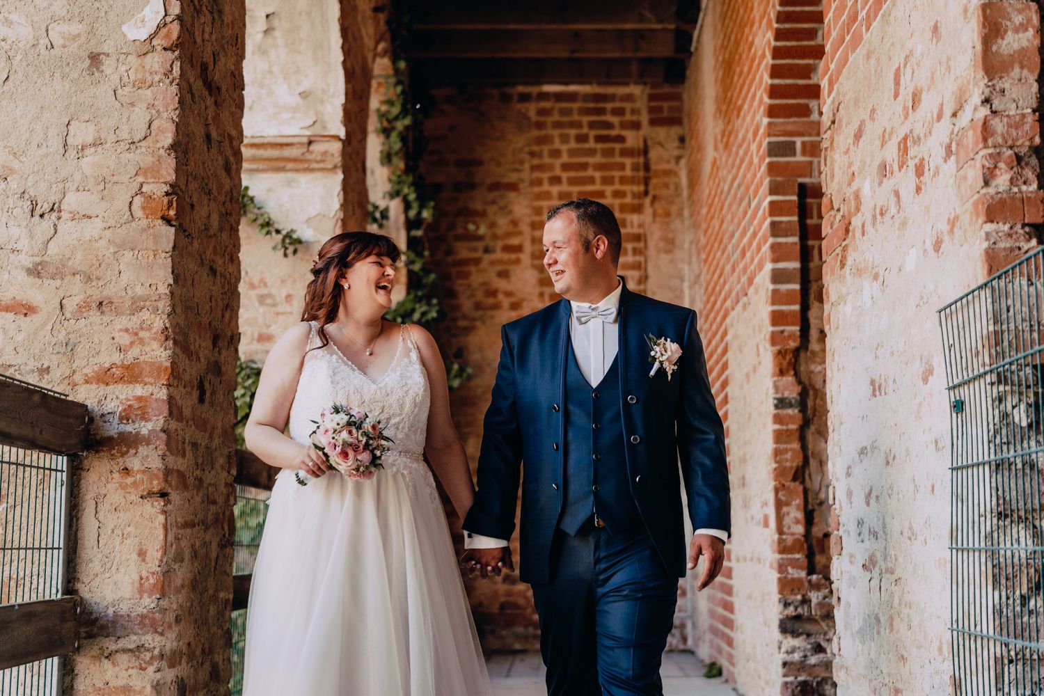 A bride and groom walk together through a rustic brick archway during their wedding day photoshoot.