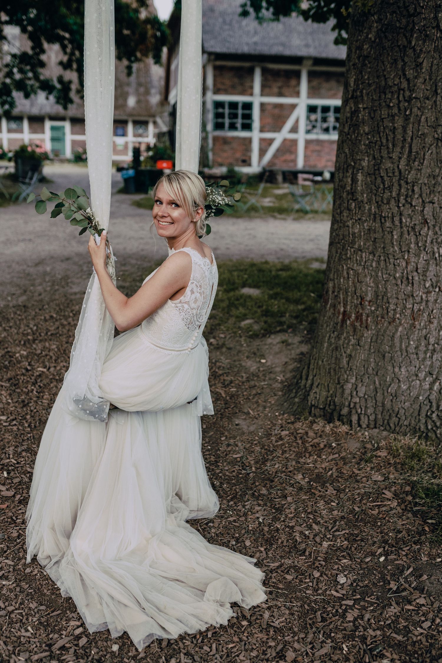 A bride in a flowing white wedding dress smiles joyfully while swinging outdoors among trees.