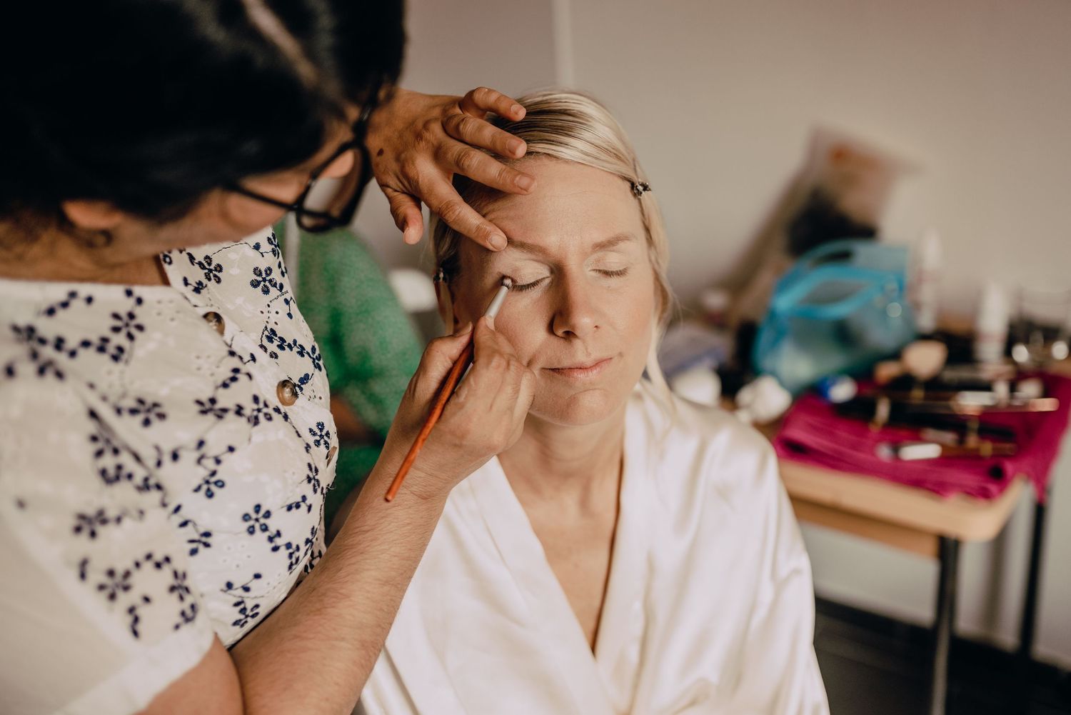 A wedding preparation scene showing final makeup touches being applied in natural lighting.