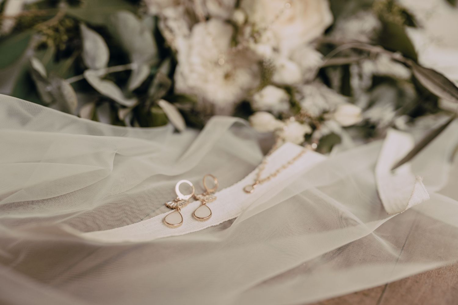 Gold wedding rings rest on delicate white tulle fabric with soft blurred flowers in the background.
