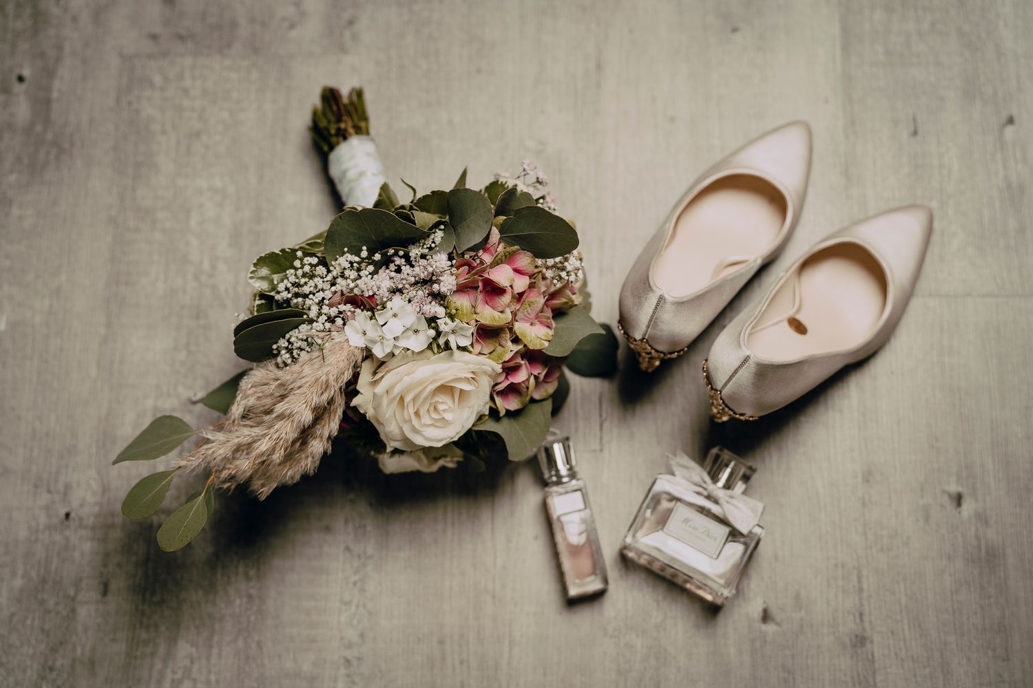 Wedding details with ivory bridal shoes, perfume bottles and a romantic blush and cream floral bouquet on grey surface.