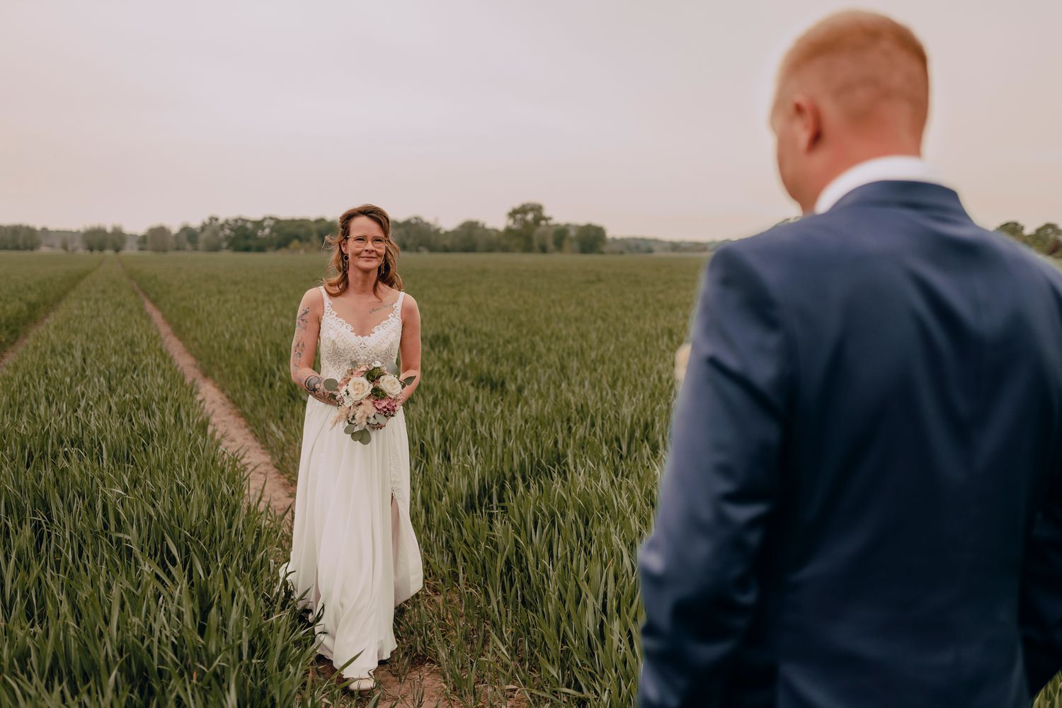 A romantic wedding moment captured in a green wheat field as bride in white dress holds bouquet.