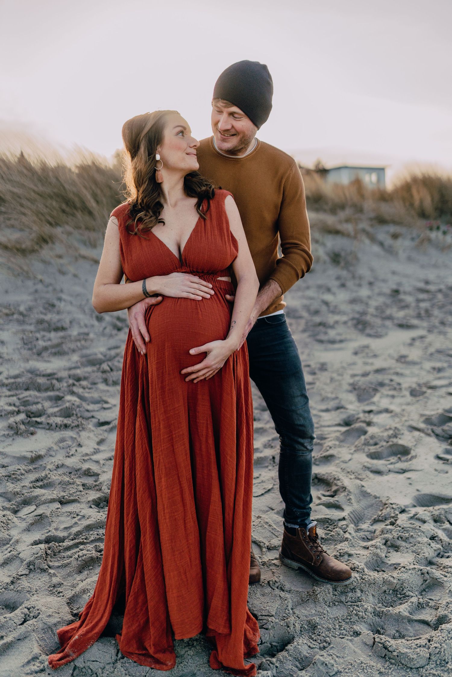 Expectant couple shares tender moments on a beach while wearing a flowing red dress and brown sweater at sunset.