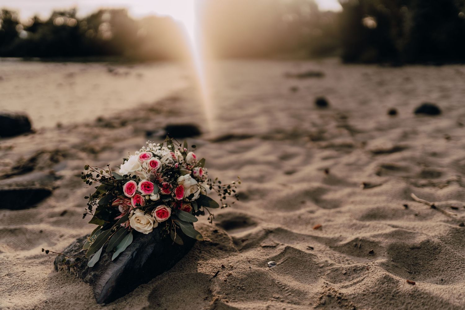 Flower bouquet with pink roses and greenery resting on sandy beach at sunset.