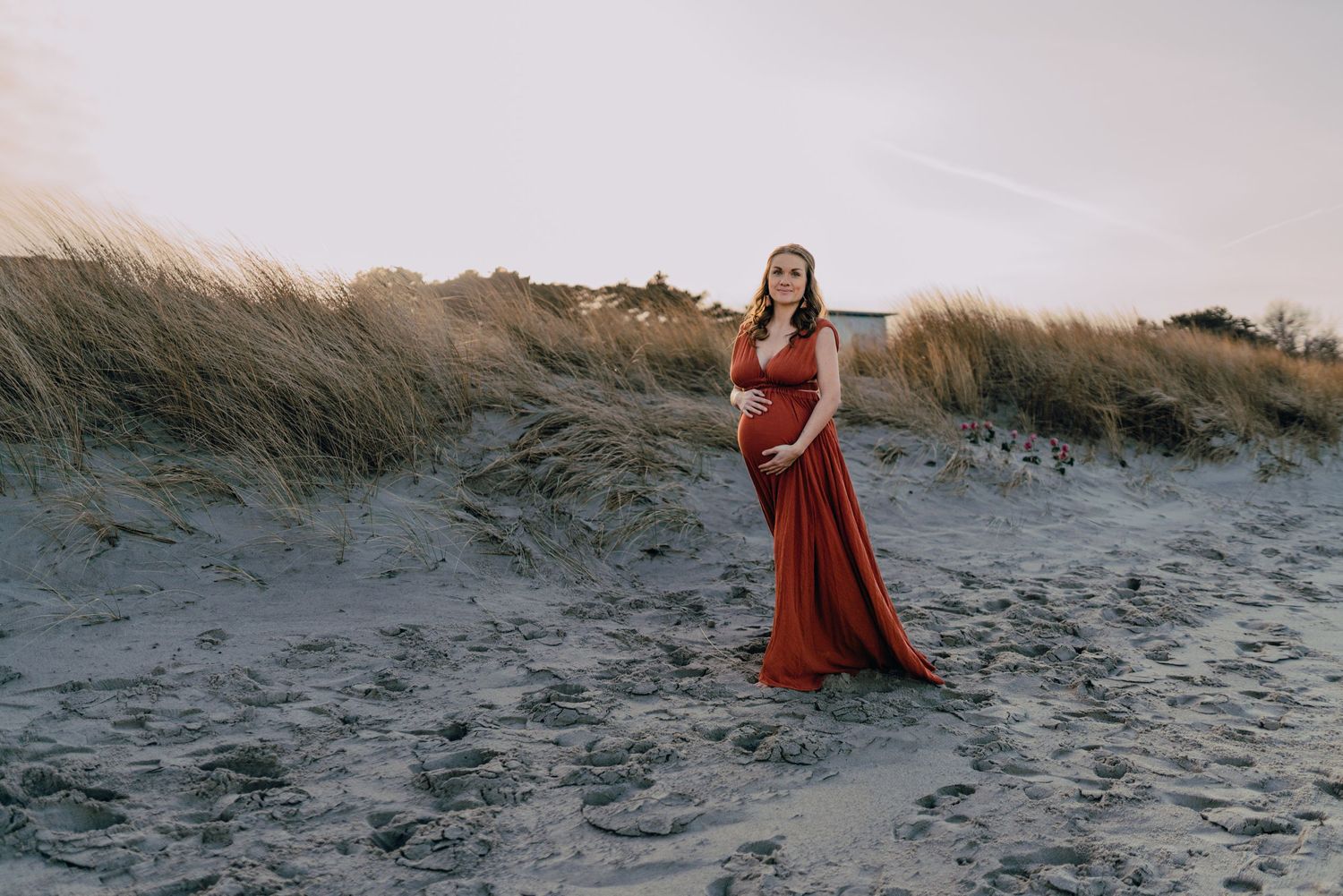 A figure in a flowing red dress walks through windswept dunes at sunset along a coastal beach landscape.