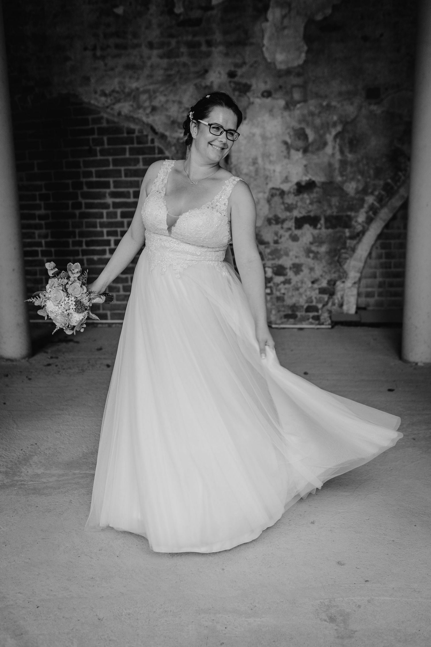 Bride in glasses twirling in a white wedding dress against exposed brick walls.