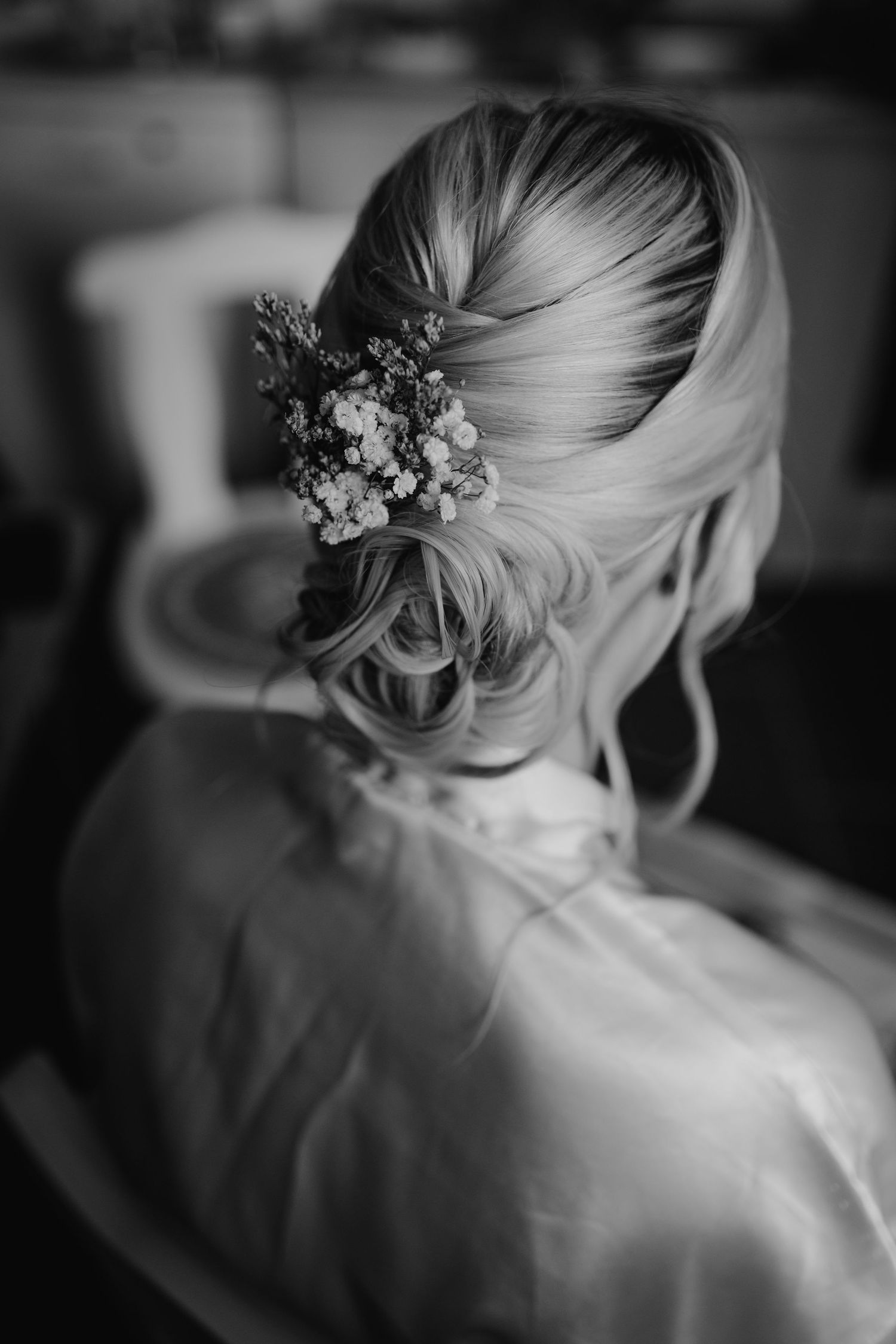 Black and white photo of an elegant bridal updo hairstyle adorned with small flowers and wispy tendrils.