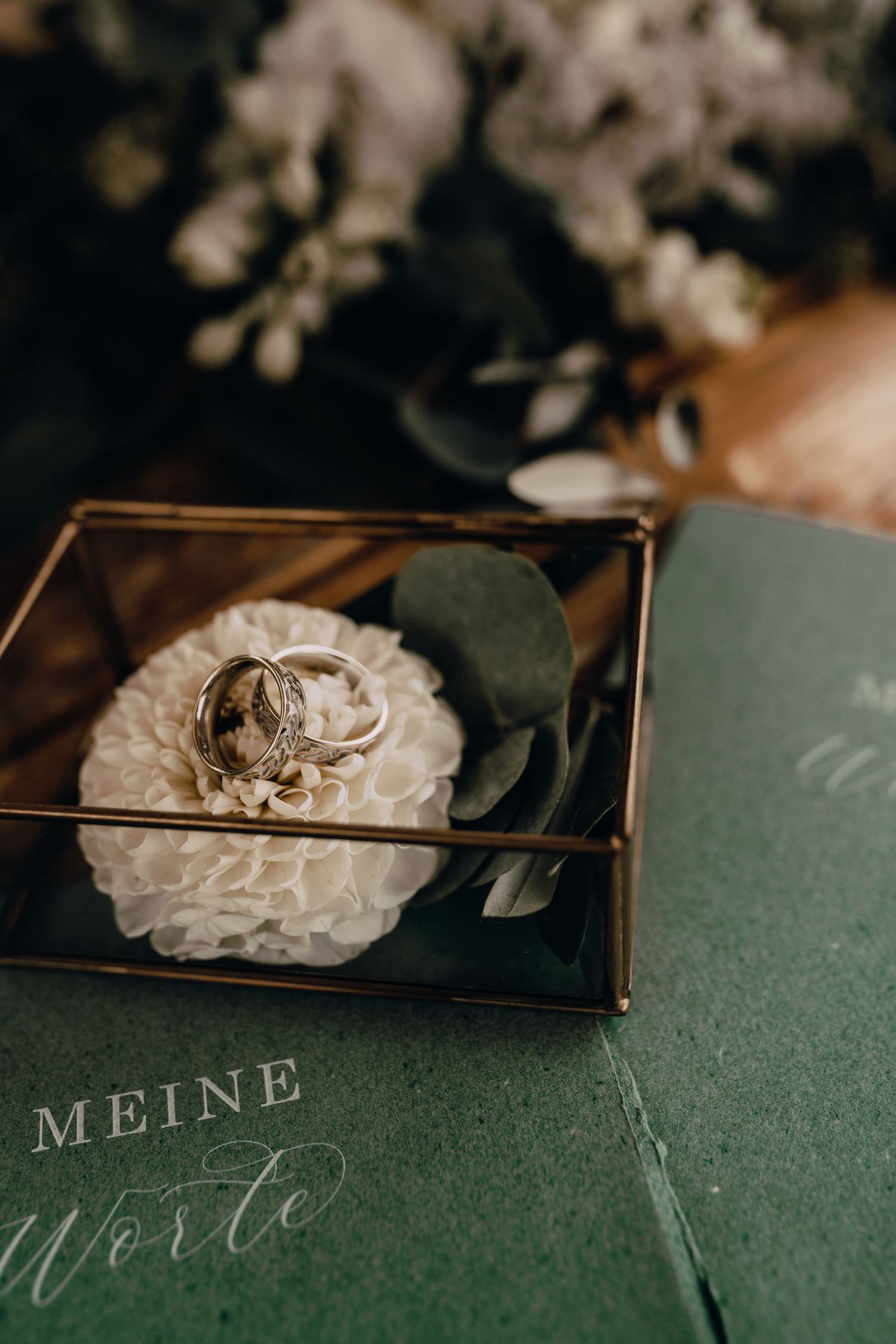 Wedding rings nestled on white flower in glass box with green book titled 'Meine' in moody lighting.
