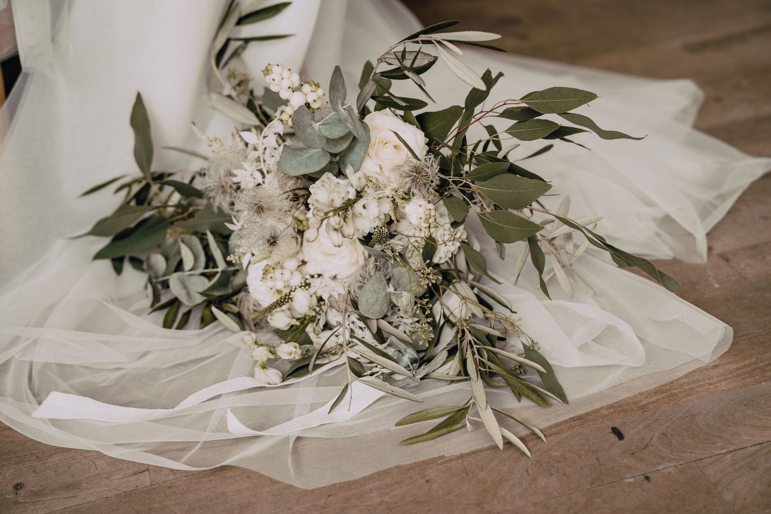 A delicate white bridal bouquet with eucalyptus leaves rests on flowing sheer fabric against a wooden floor.