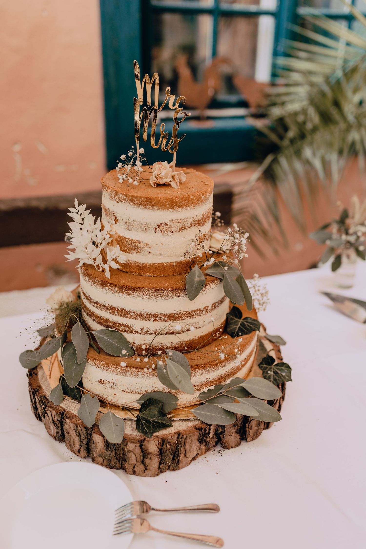 A rustic naked wedding cake decorated with eucalyptus leaves sits on a wood slice cake stand on a white tablecloth.