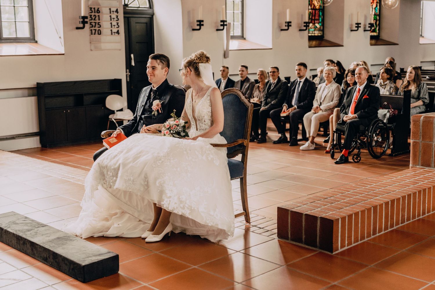 A couple seated during a church wedding ceremony with guests attending in the background.